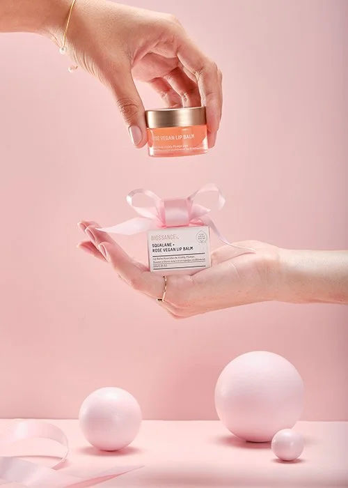 Two hands exchanging a small jar of vegan lip balm and a small box of rose vegan lip balm against a light pink background with decorative pink and white spheres.