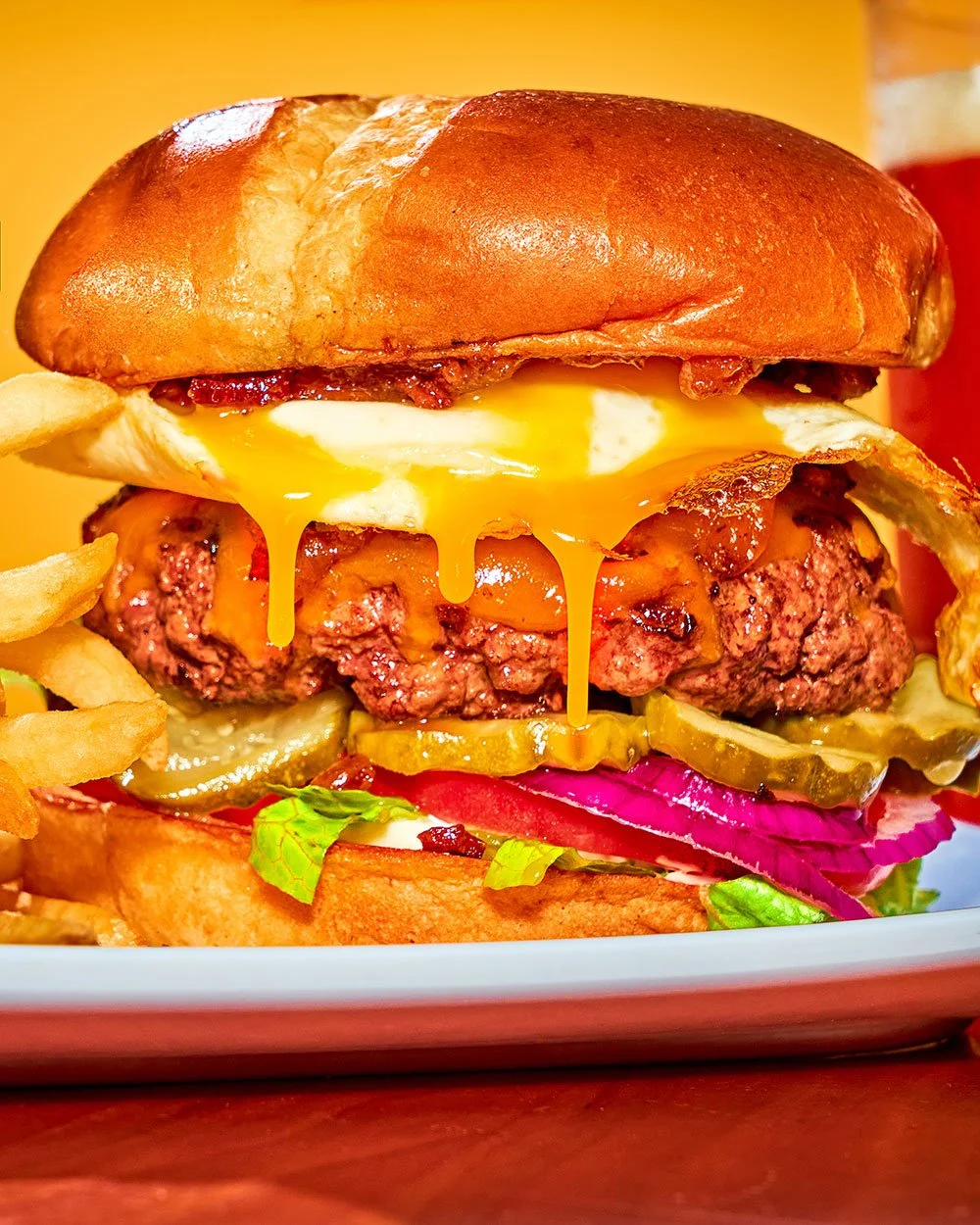 Close-up of a cheeseburger with a fried egg, lettuce, tomato, pickles, onions, bacon, and melted cheese, served with French fries on a tray.