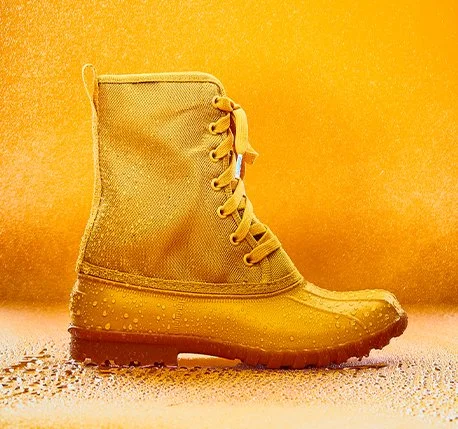 A yellow rain boot with a textured upper, laces on the side, and water droplets on the sole, set against a yellow background.