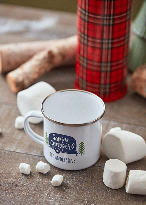 A white mug with a silver rim and a "happy campers" label, surrounded by marshmallows and decorative logs on a wooden table.