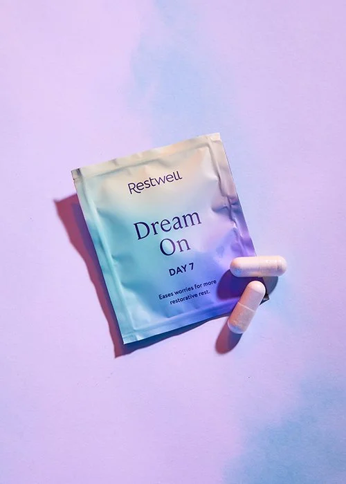 A pastel-colored packet labeled 'Restwell Dream On Day 7' with two white capsules beside it on a purple background.