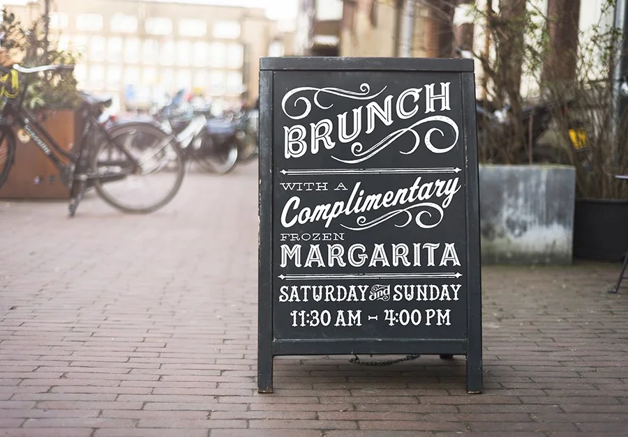 A black sidewalk sign advertising brunch with a complimentary frozen margarita on Saturdays and Sundays from 11:30 am to 4:00 pm, placed on a brick sidewalk outdoors.