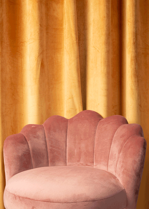 A pink velvet chair with a scalloped backrest in front of gold curtains.