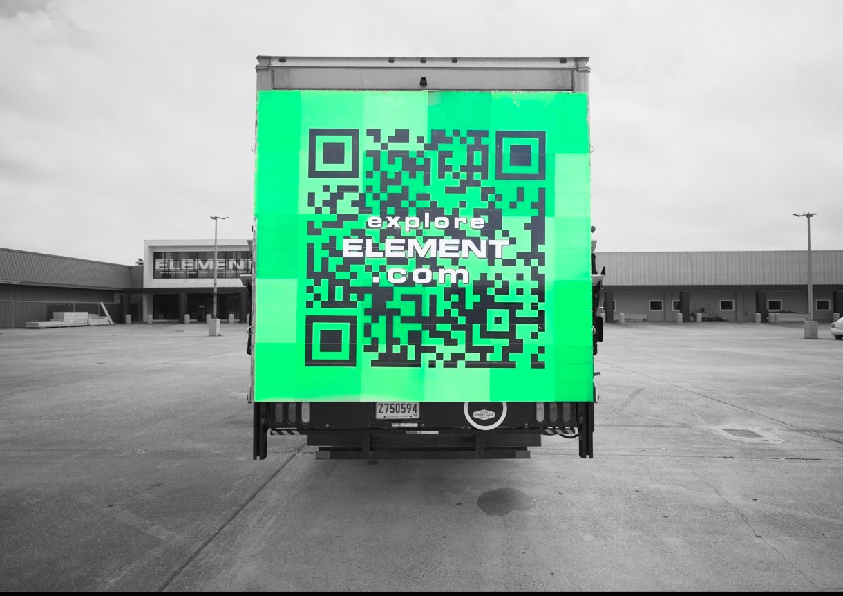 A truck with a green digital sign displaying a QR code and the website explore ELEMENT .com, parked in an empty parking lot outside a strip mall with a cloudy sky.
