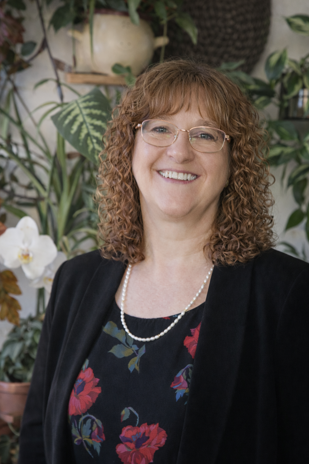 A woman with curly red hair and glasses smiling, wearing a black blazer and a floral blouse with a pearl necklace, standing in front of a background of green plants and white orchids.