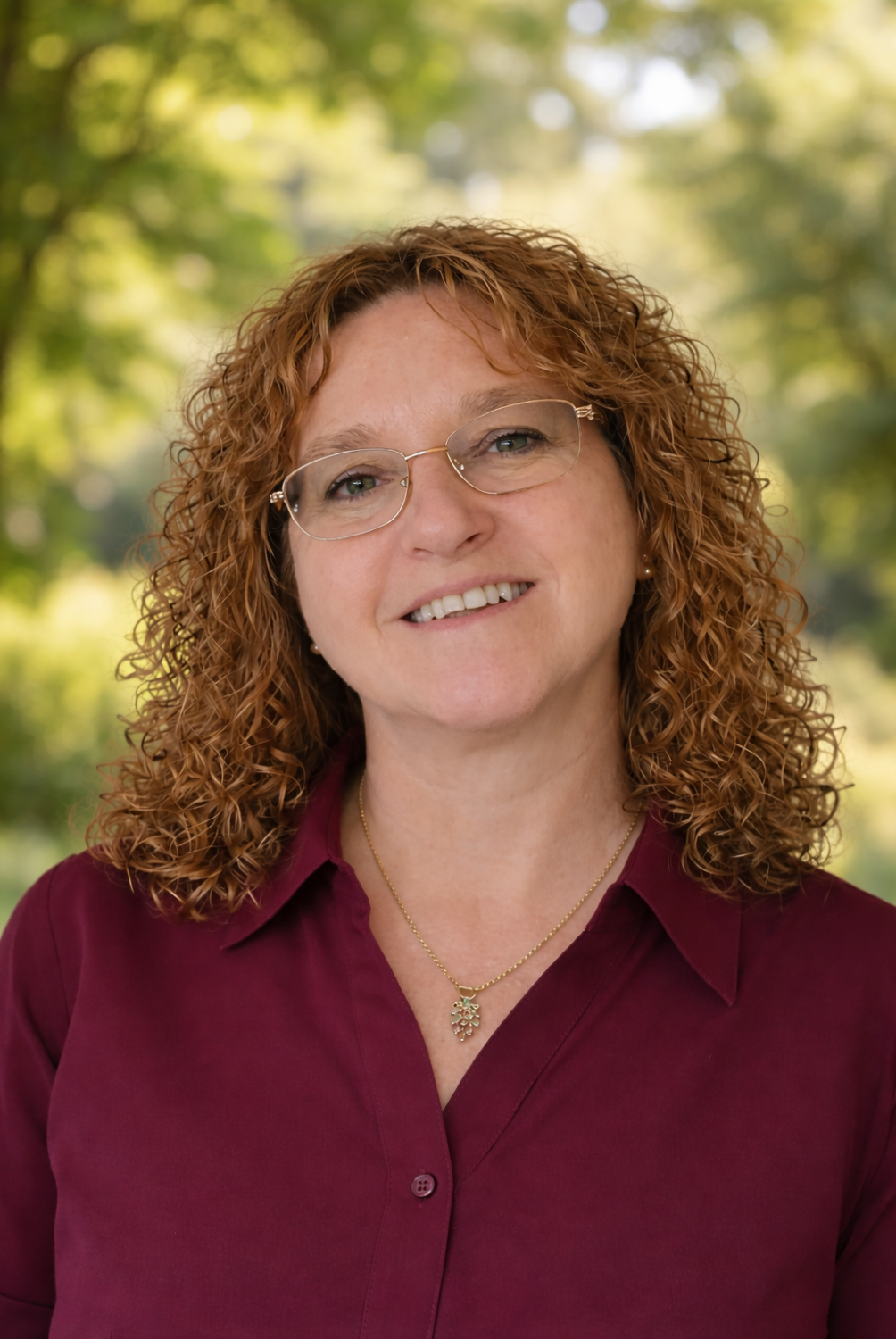 A woman with curly red hair and glasses smiling outdoors with a blurred green and yellow background.