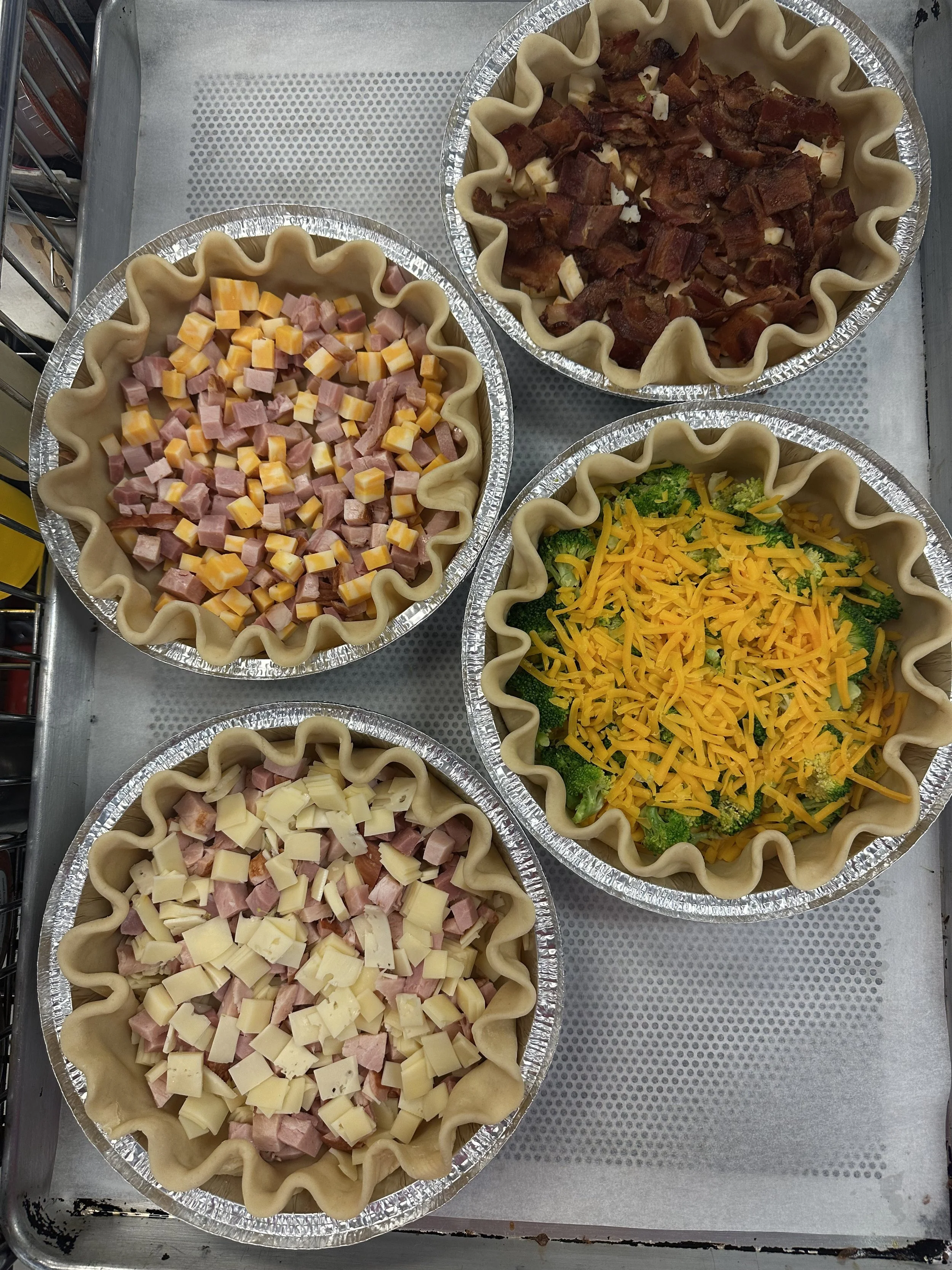 Four aluminum pie dishes filled with layers of ingredients for savory pies, including ham, cheese, broccoli, bacon, and shredded cheddar cheese.
