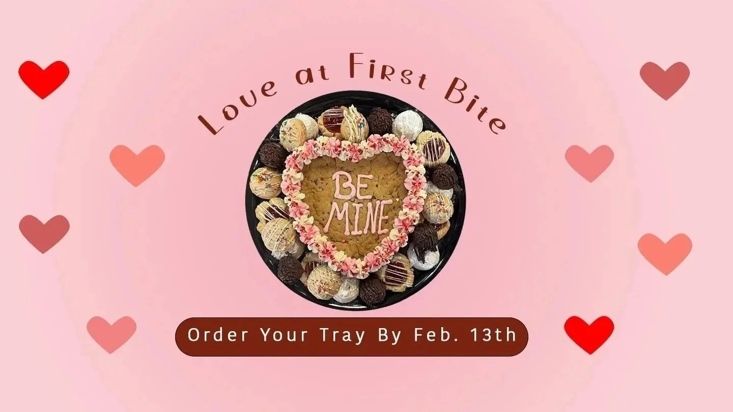Valentine's Day-themed image featuring a heart-shaped cookie with pink icing and the words 'Be Mine' surrounded by assorted chocolates and cookies on a black tray. Pink and red hearts decorate the background. Text says 'Love at First Bite' and 'Order