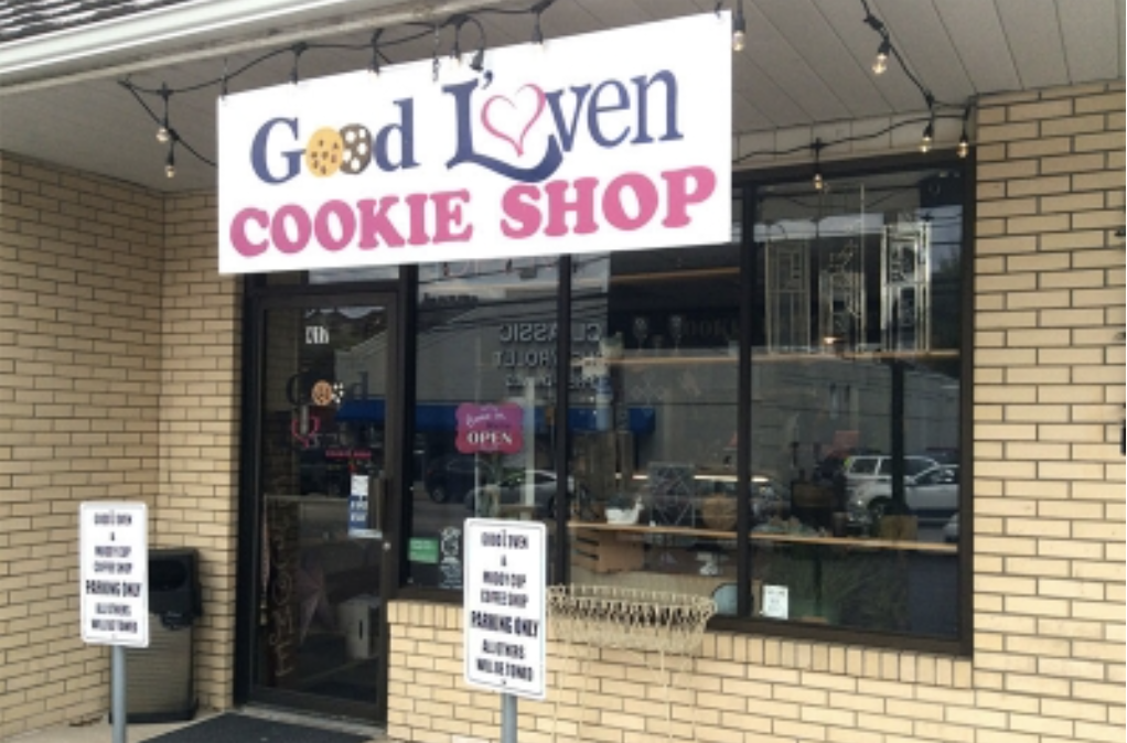 Exterior view of a cookie shop called 'Good Lovin Cookie Shop' with a sign overhead, a glass door, and parking signs in front.