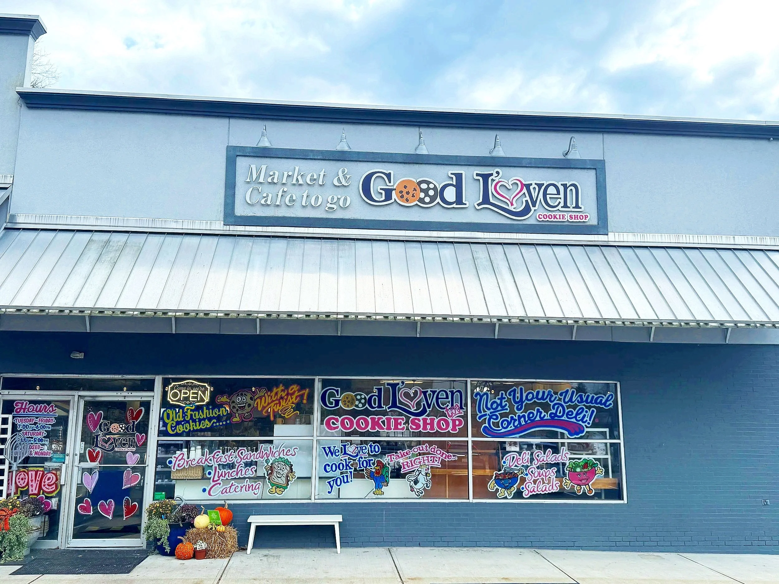 Front of a bakery and cafe called 'Good L'Oven' with colorful window signs advertising cookies, sandwiches, salads, and catering, decorated with hearts and cartoons, pumpkins outside, and a sign stating it is open.