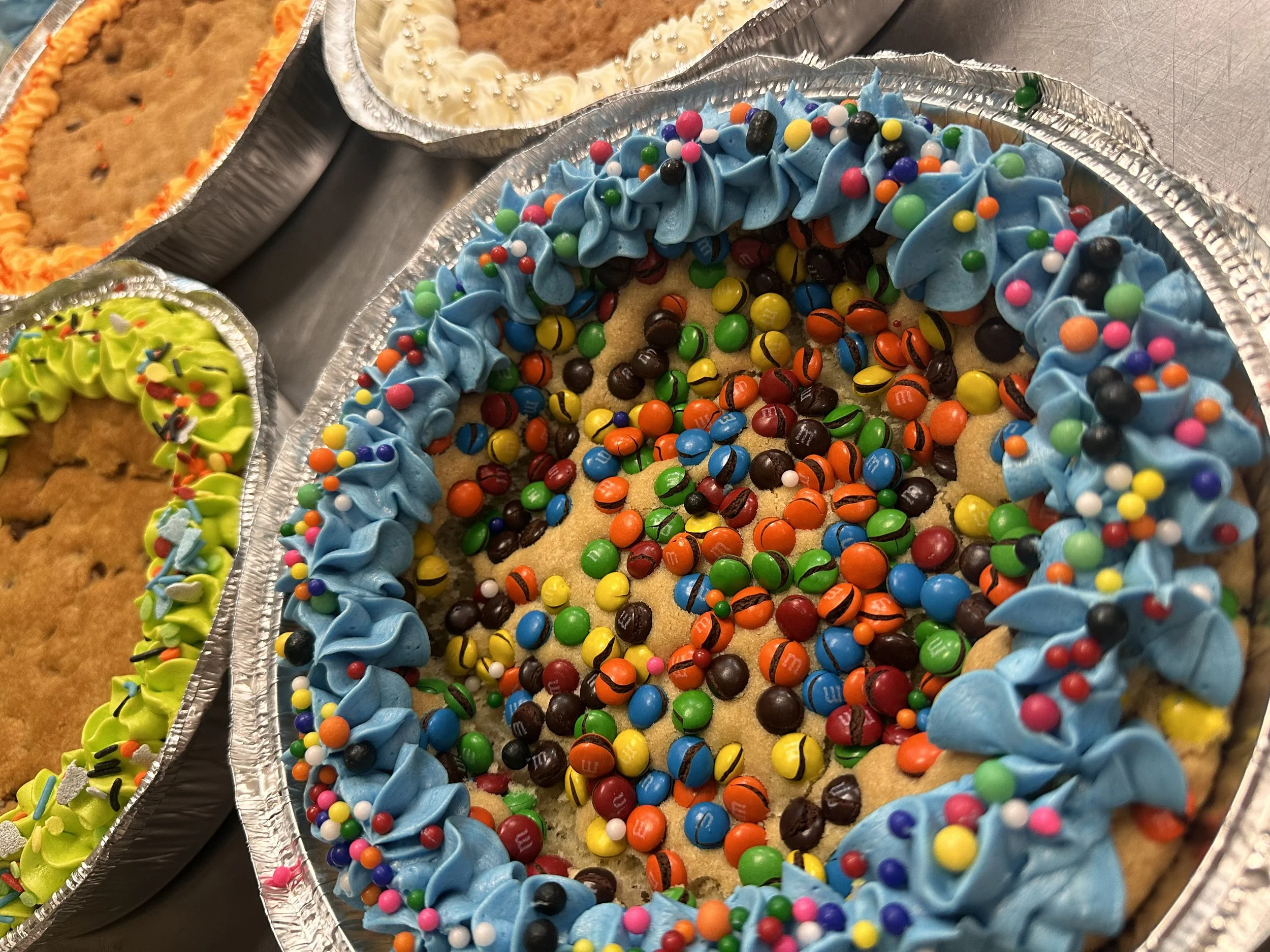 Decorated cookies in aluminum pans, including one with blue icing and colorful sprinkles, and others with yellow and white icing.