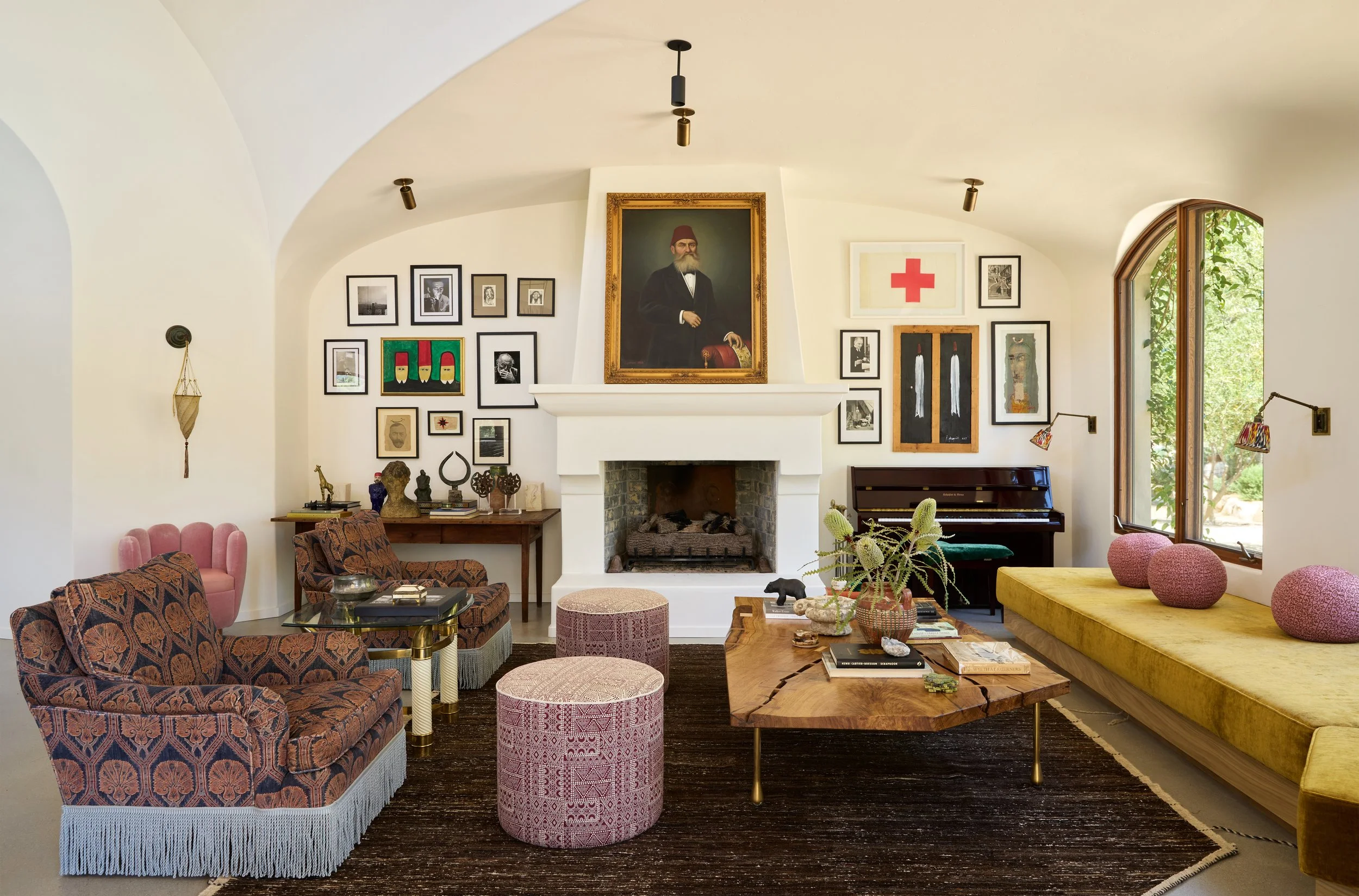 Living room with a white fireplace, eclectic art on the walls, a pink cushioned armchair, patterned sofas, a wooden coffee table with books and decor, a green window seat with pink cushions, and various decorative items on furniture.
