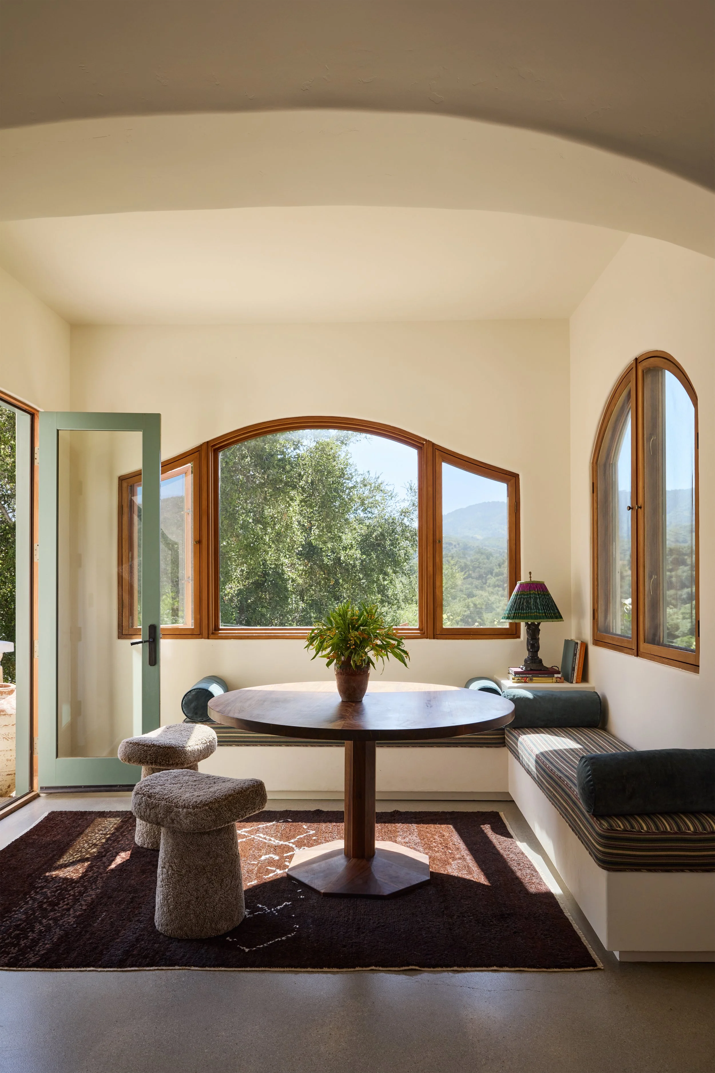Sunlit dining nook with a round wooden table, a potted plant, two plush stool chairs, built-in bench seating with striped cushions, and large arched windows overlooking trees and mountains.