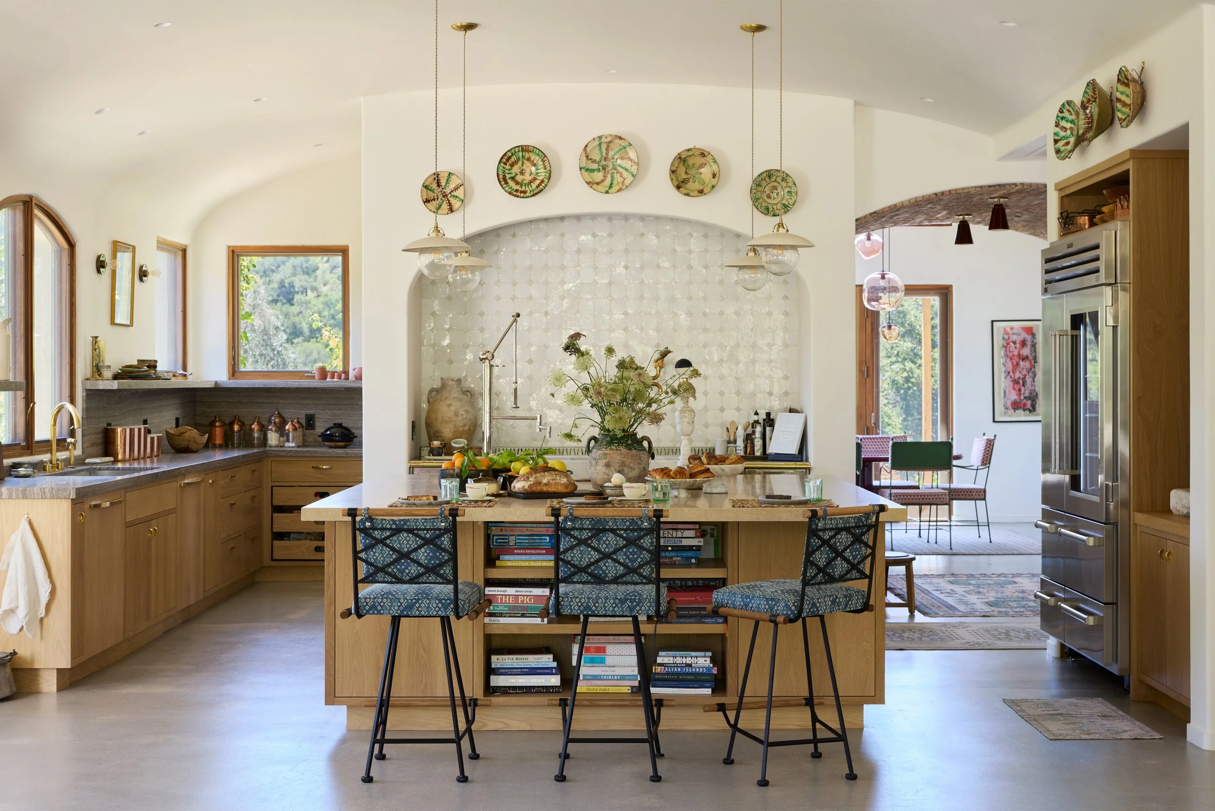 A modern kitchen with a central island, wooden cabinets, and various decorative plates on the wall, with a dining area visible in the background.