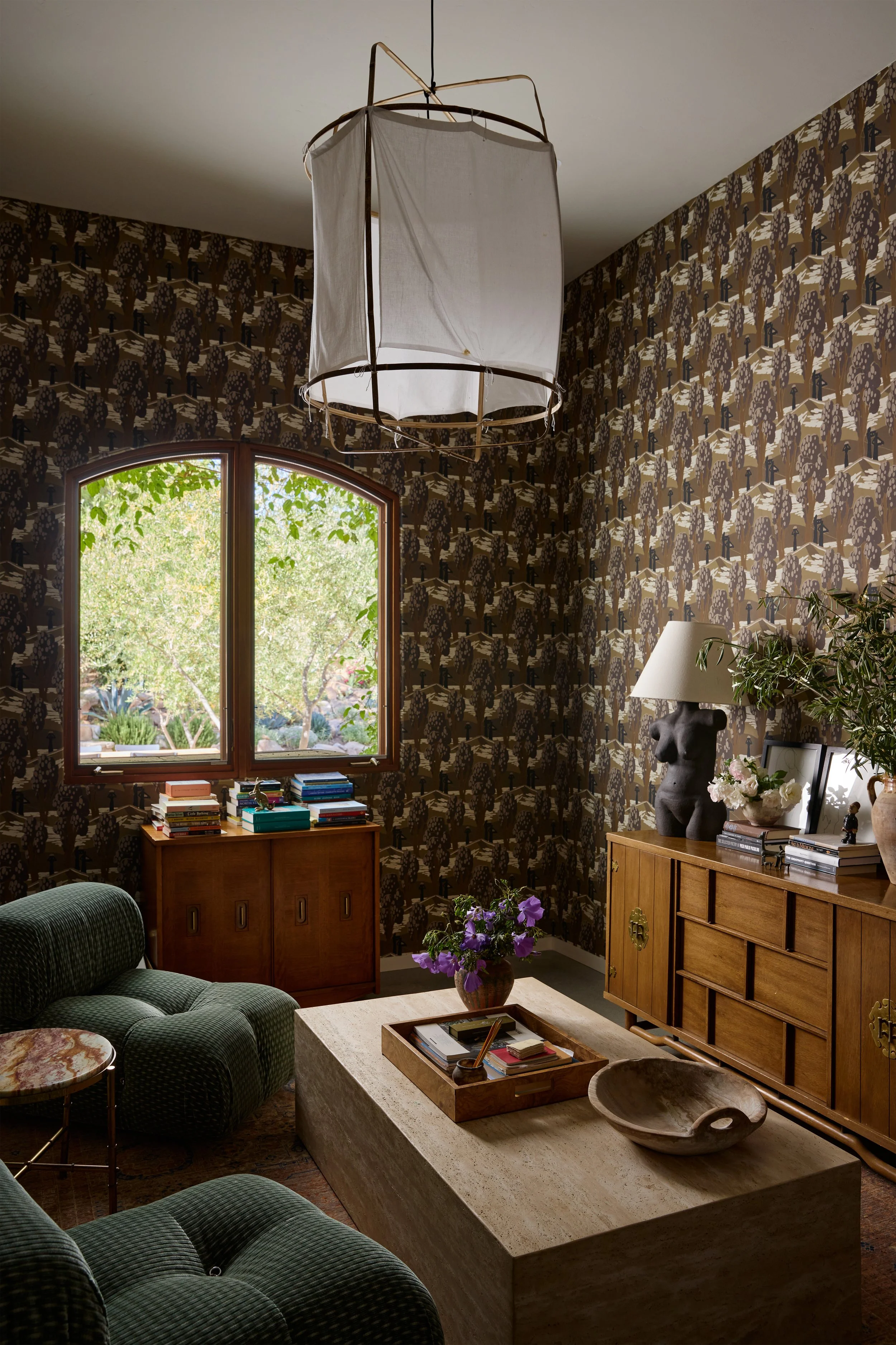 A cozy living room with a patterned wallpaper, a large window showing greenery outside, green upholstered chairs, a stone coffee table with decorative items, a wooden sideboard with books and a lamp, and a black torso-shaped sculpture used as a lamp base.