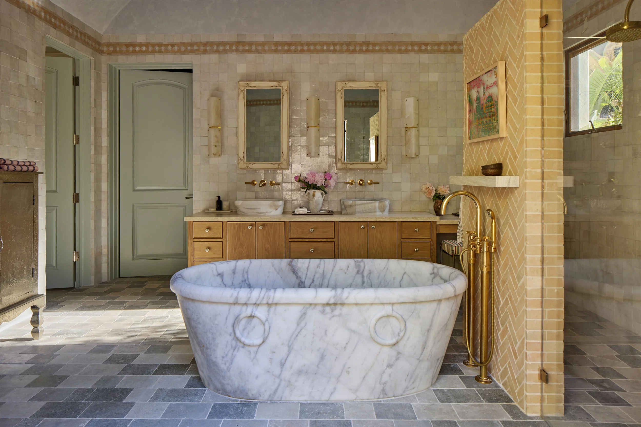 Modern bathroom with a marble bathtub, wooden vanity with two vessel sinks, large mirrors, and floral decorations. There are windows providing natural light to the space.