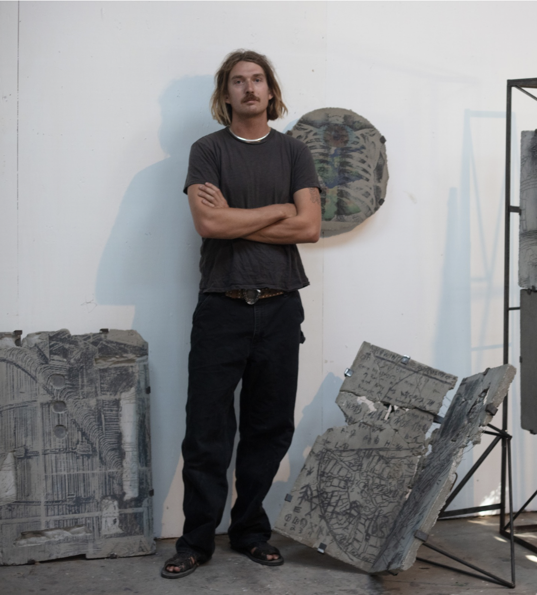 A young man with shoulder-length hair and a mustache stands with arms crossed in an art studio surrounded by abstract concrete sculptures and artworks on the wall.