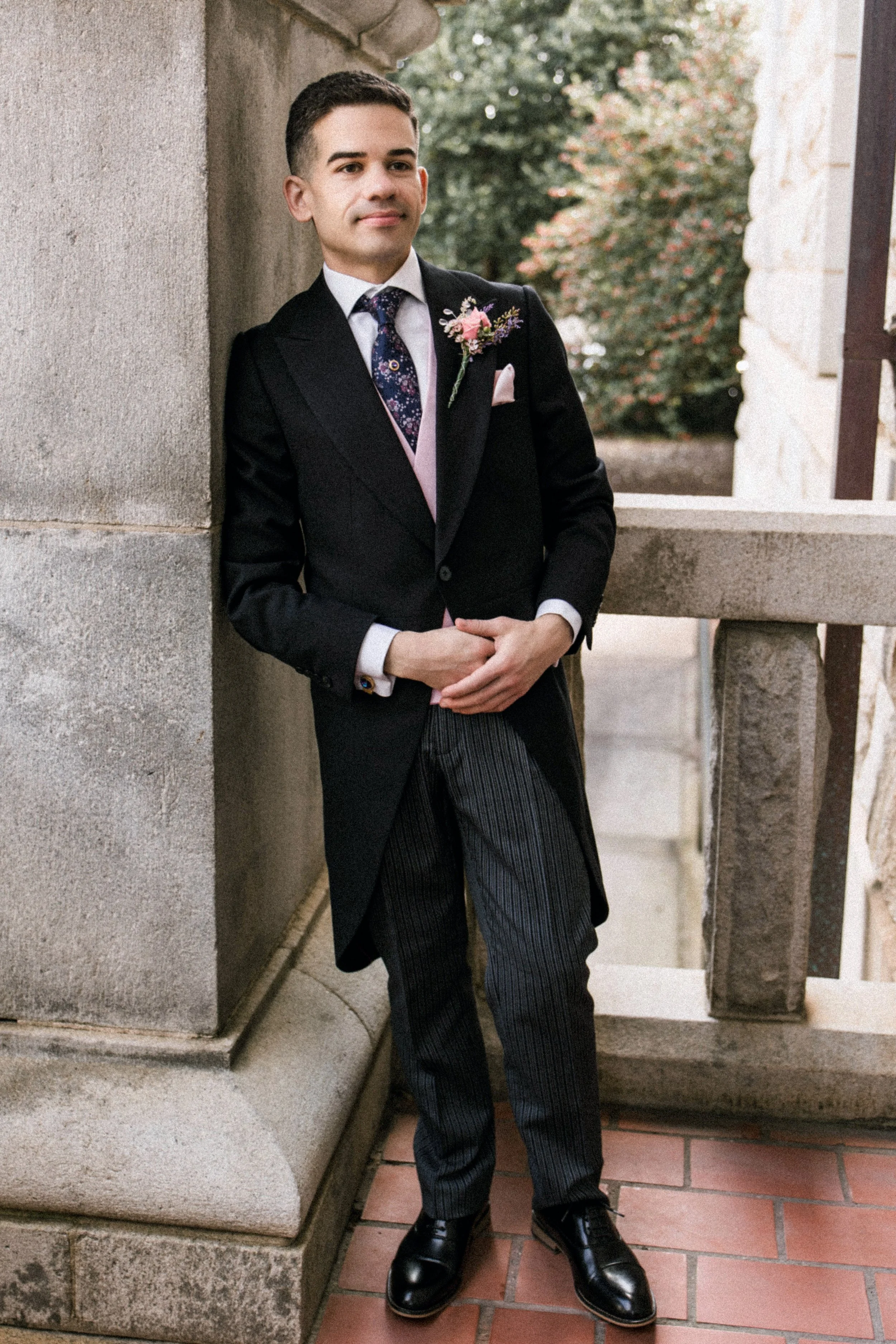 A young man dressed in a black tuxedo with a floral boutonniere, standing outdoors on a brick patio next to a stone building, looking confidently at the camera.