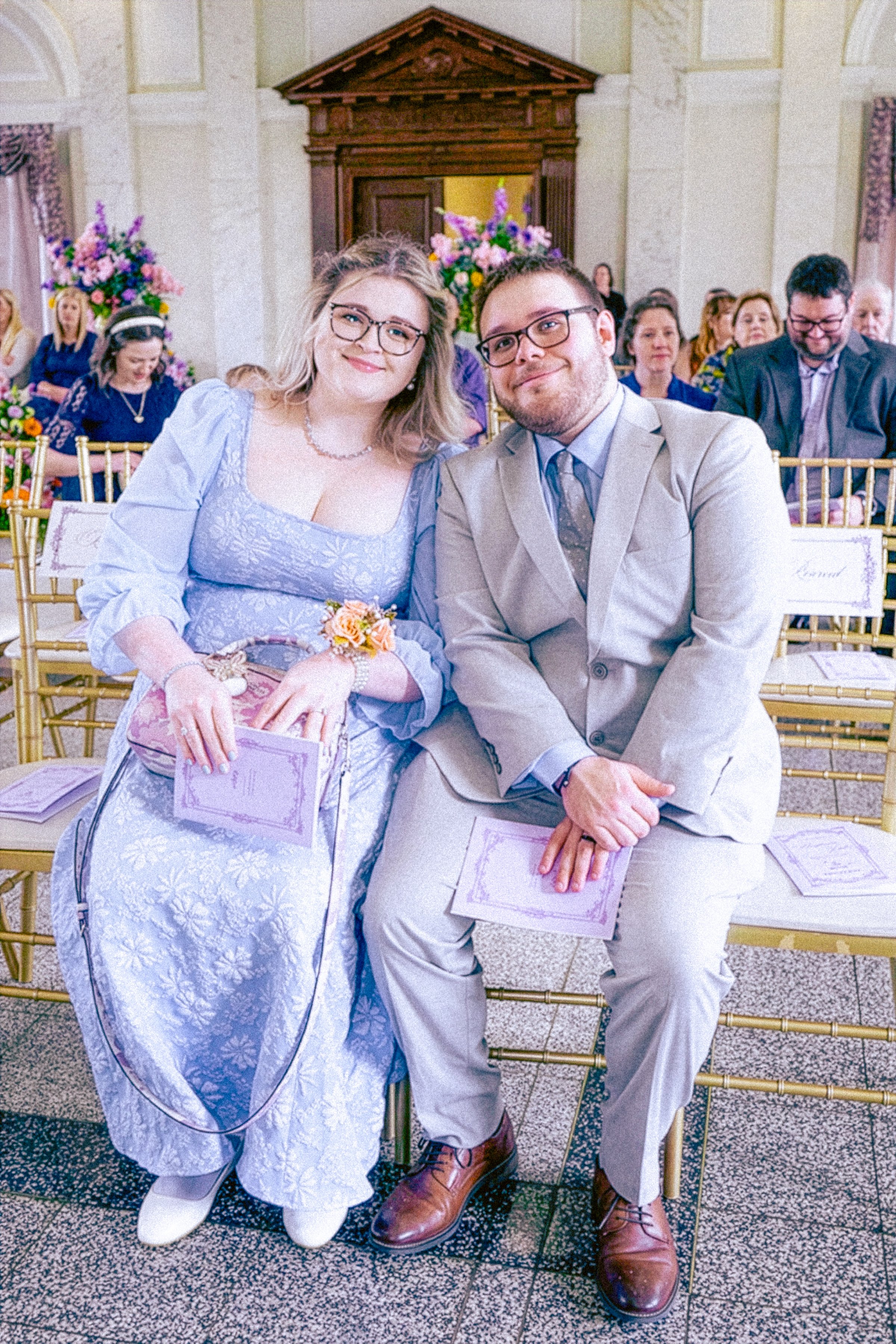 A couple sitting at a wedding ceremony, holding programs, with smiling expressions. The woman wears glasses and a light purple dress, and the man wears glasses and a beige suit. The background shows guests, floral arrangements, and an ornate wooden d