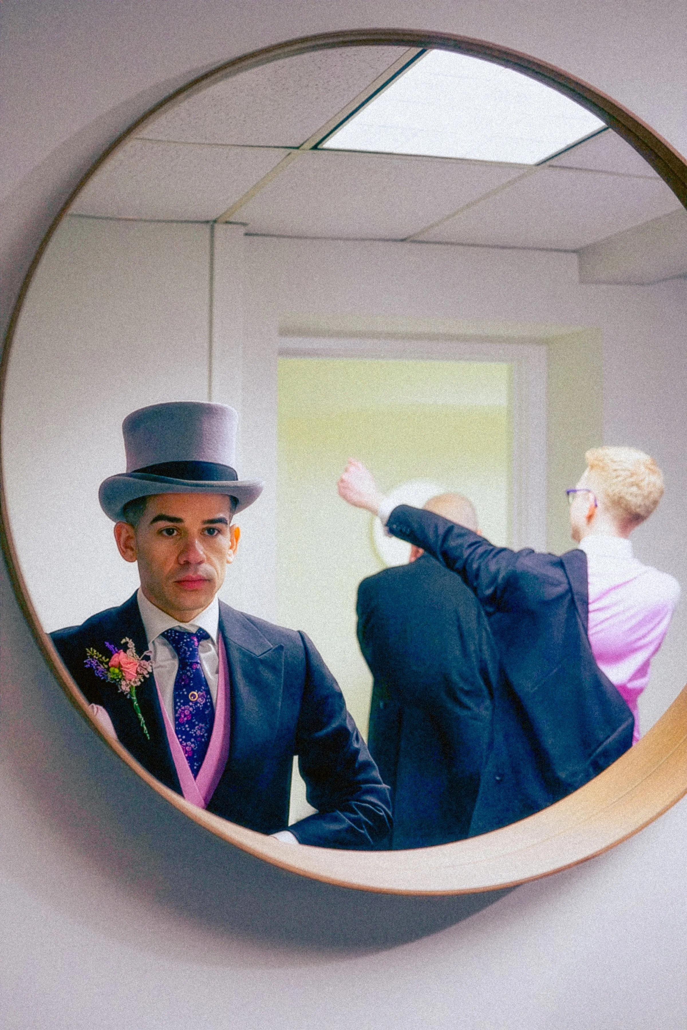 Man in a suit wearing a top hat and a boutonnière, looking at his reflection in a mirror.