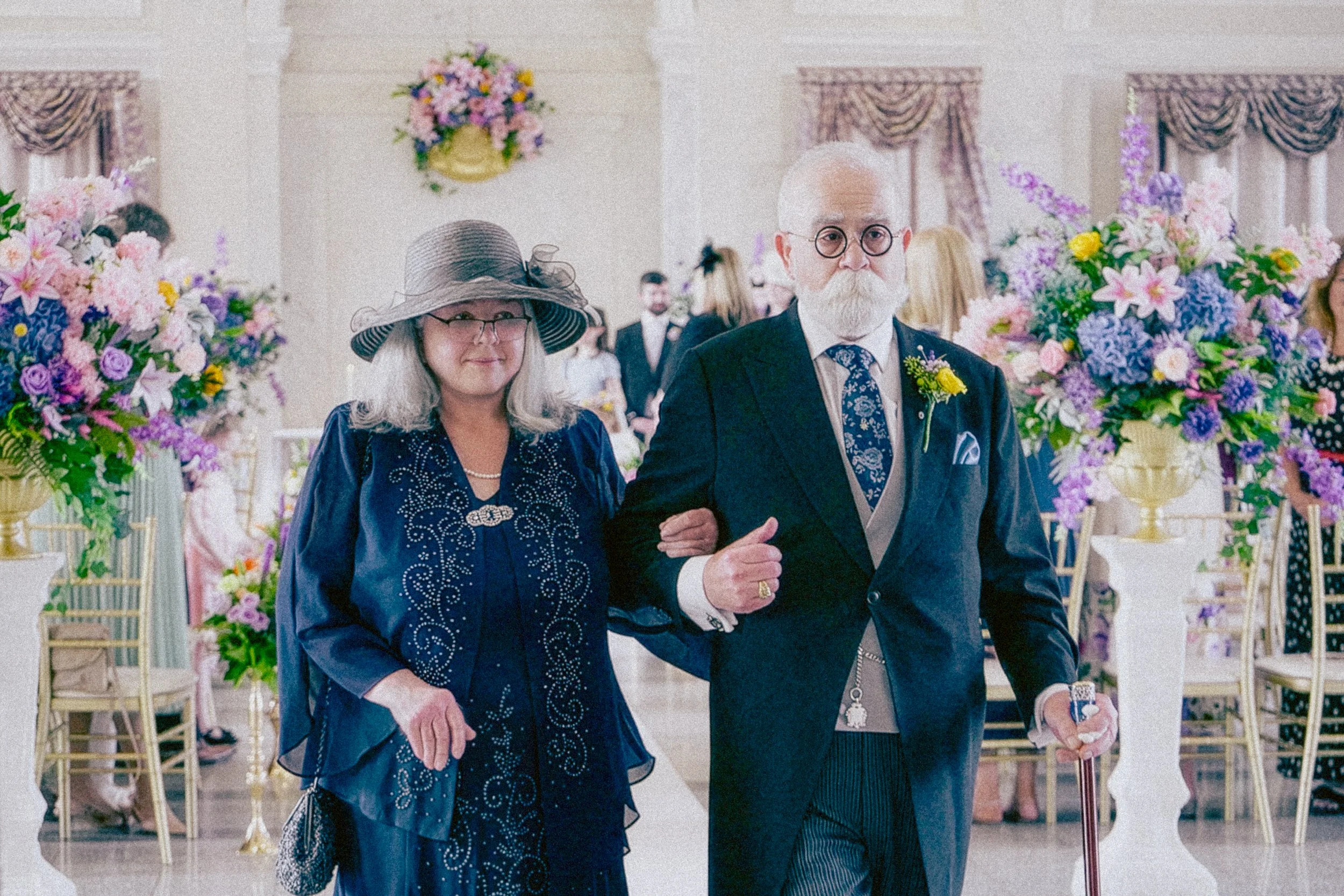 An elderly man and woman walking arm-in-arm at a wedding reception decorated with colorful flowers, gold chairs, and floral arrangements.