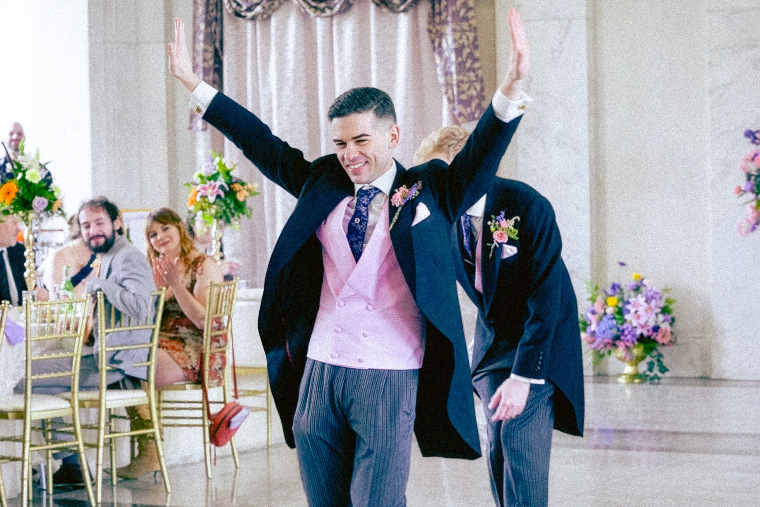 Two men in formal suits are dancing together at a wedding reception, with one man wearing a pink vest and the other in a dark suit. They are smiling and appear to be enjoying the moment. Guests are seated at decorated tables with floral arrangements,