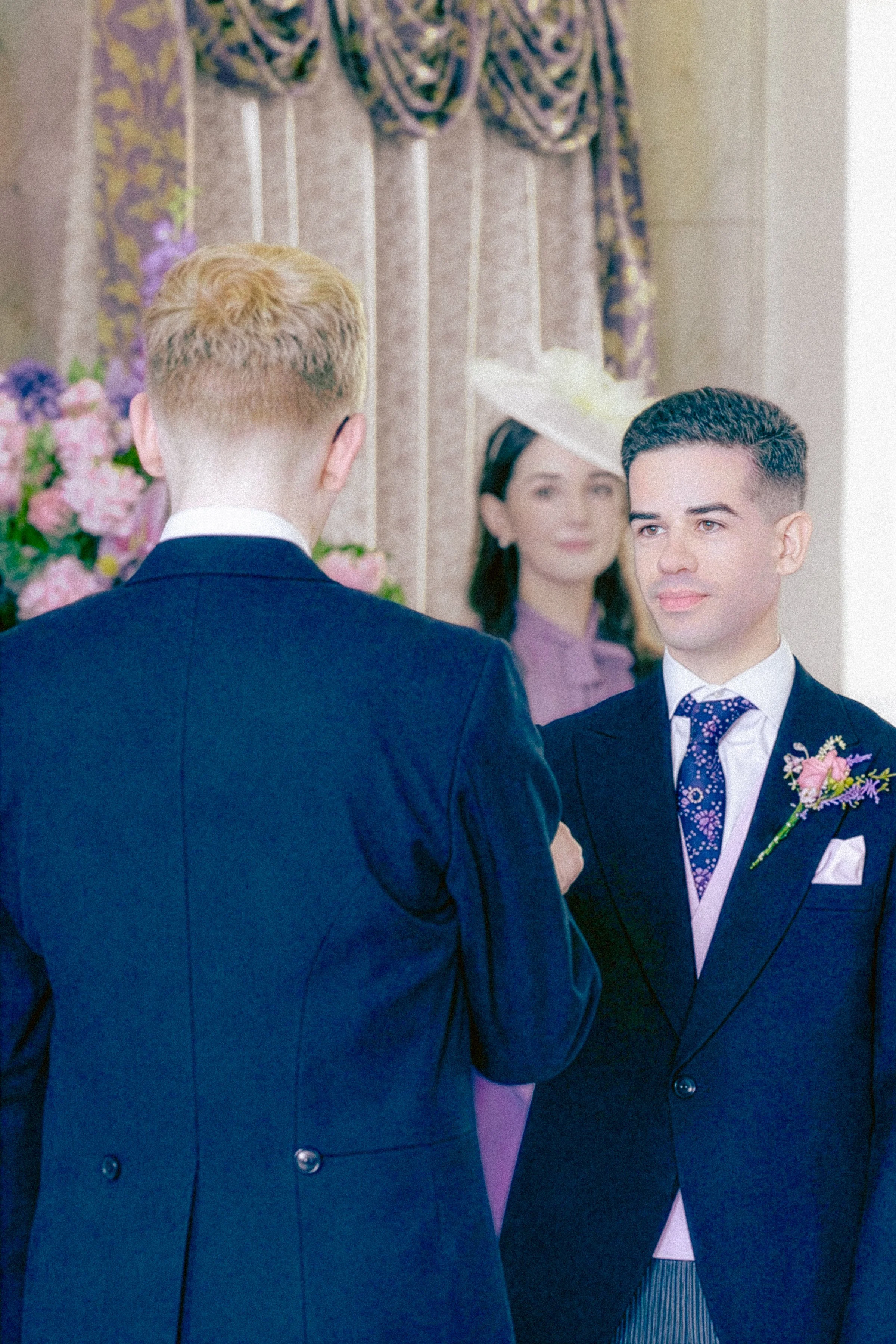 A wedding ceremony with a groom and a bride exchanging vows, with a woman in a hat in the background and floral decorations.