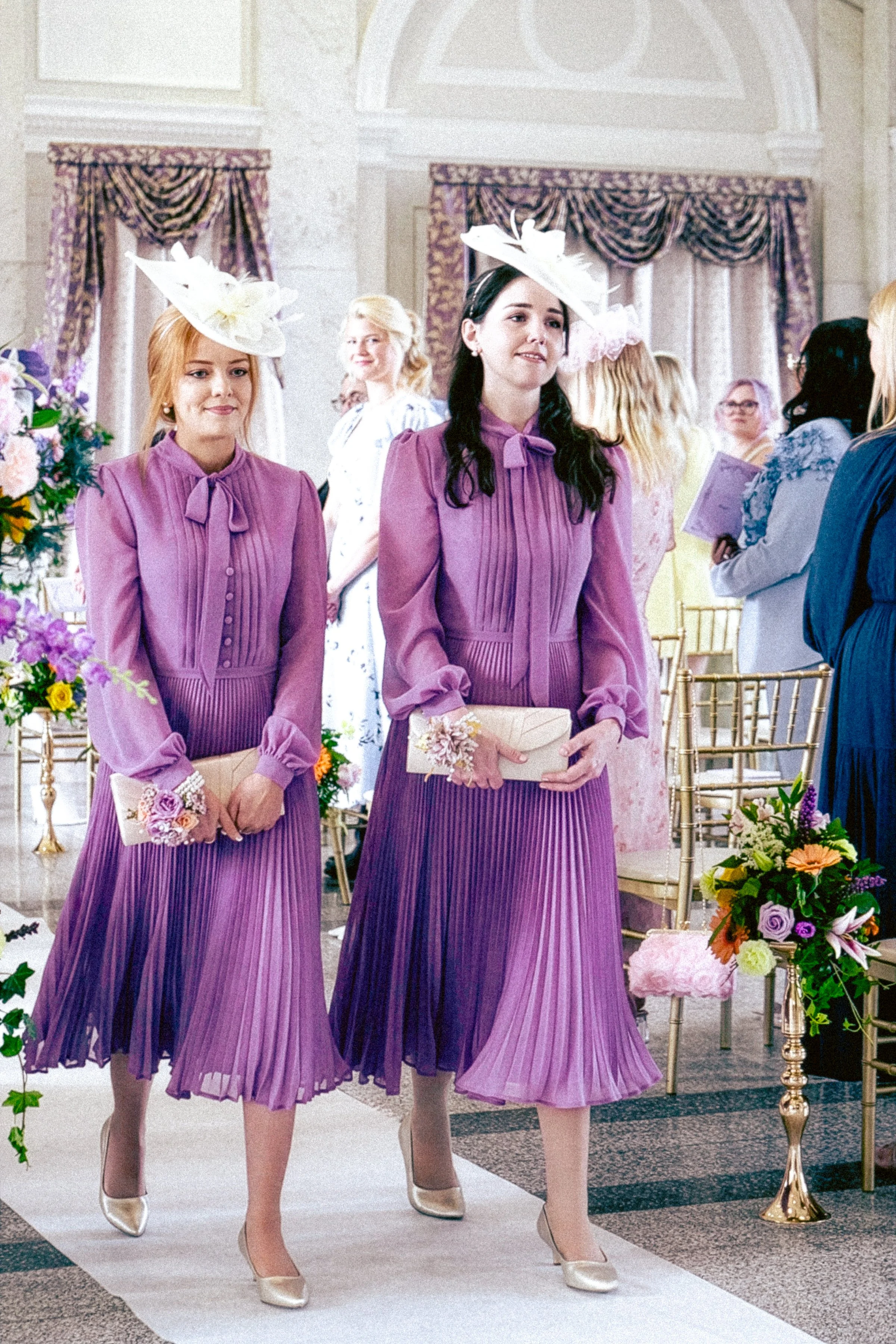 Two women in lavender pleated dresses and white hats with feathers walking down the aisle of a wedding ceremony, holding clutch purses with corsages, in an ornate room with purple curtains and floral arrangements.