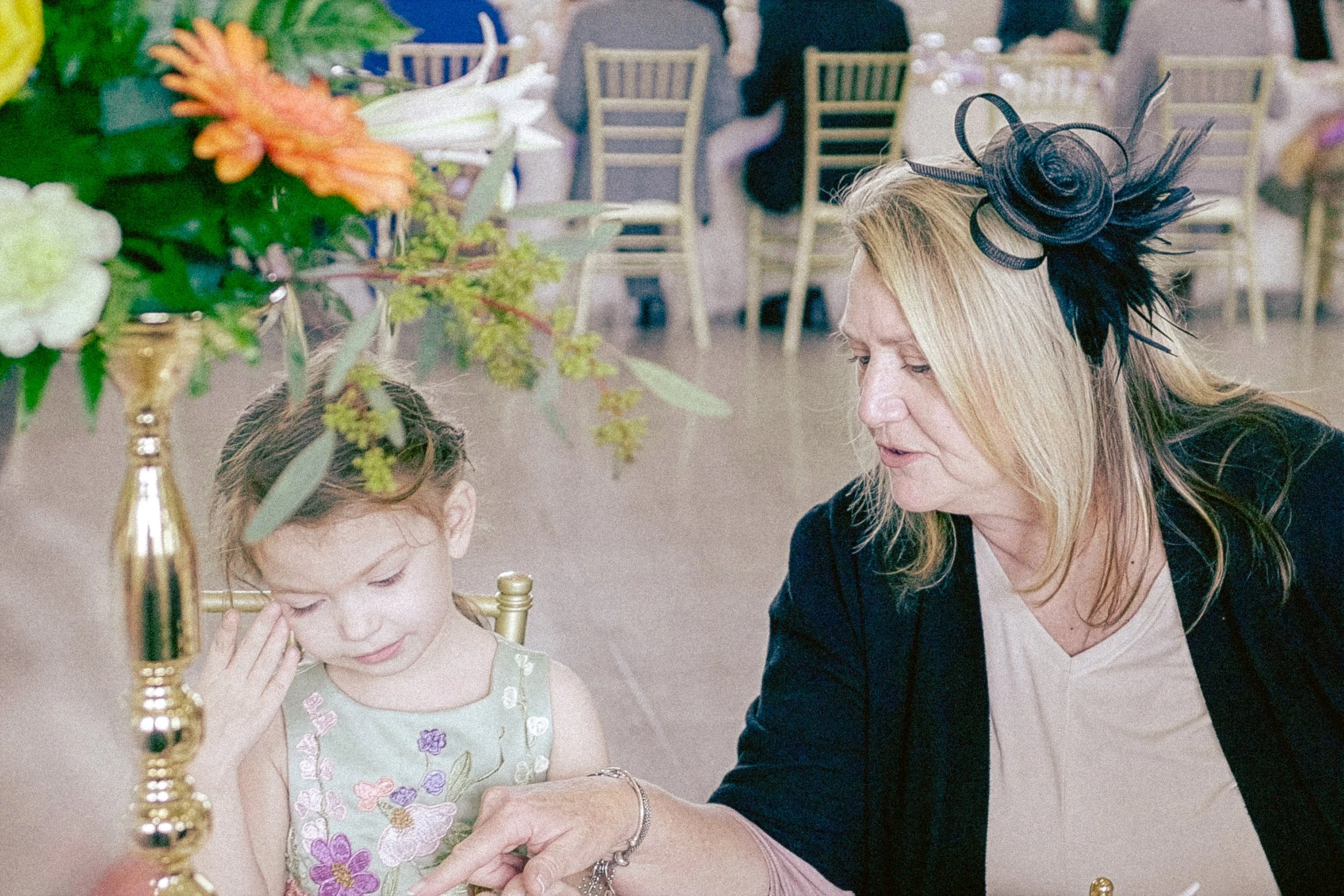 A woman in a black dress and a decorative black hat is sitting at a table with a young girl. The girl is wearing a light green dress with embroidered flowers. The woman appears to be helping the girl adjust her clothing or jewelry. The background sho