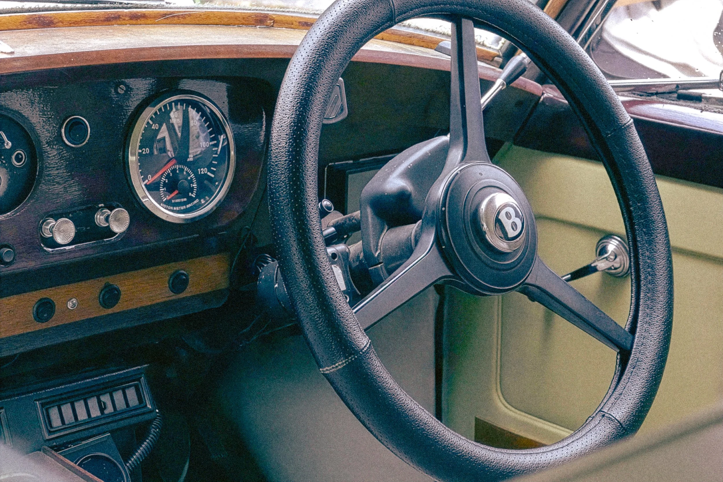 Interior of a vintage car showing the steering wheel with a Bentley logo, dashboard with speedometer, and some control knobs and gauges.