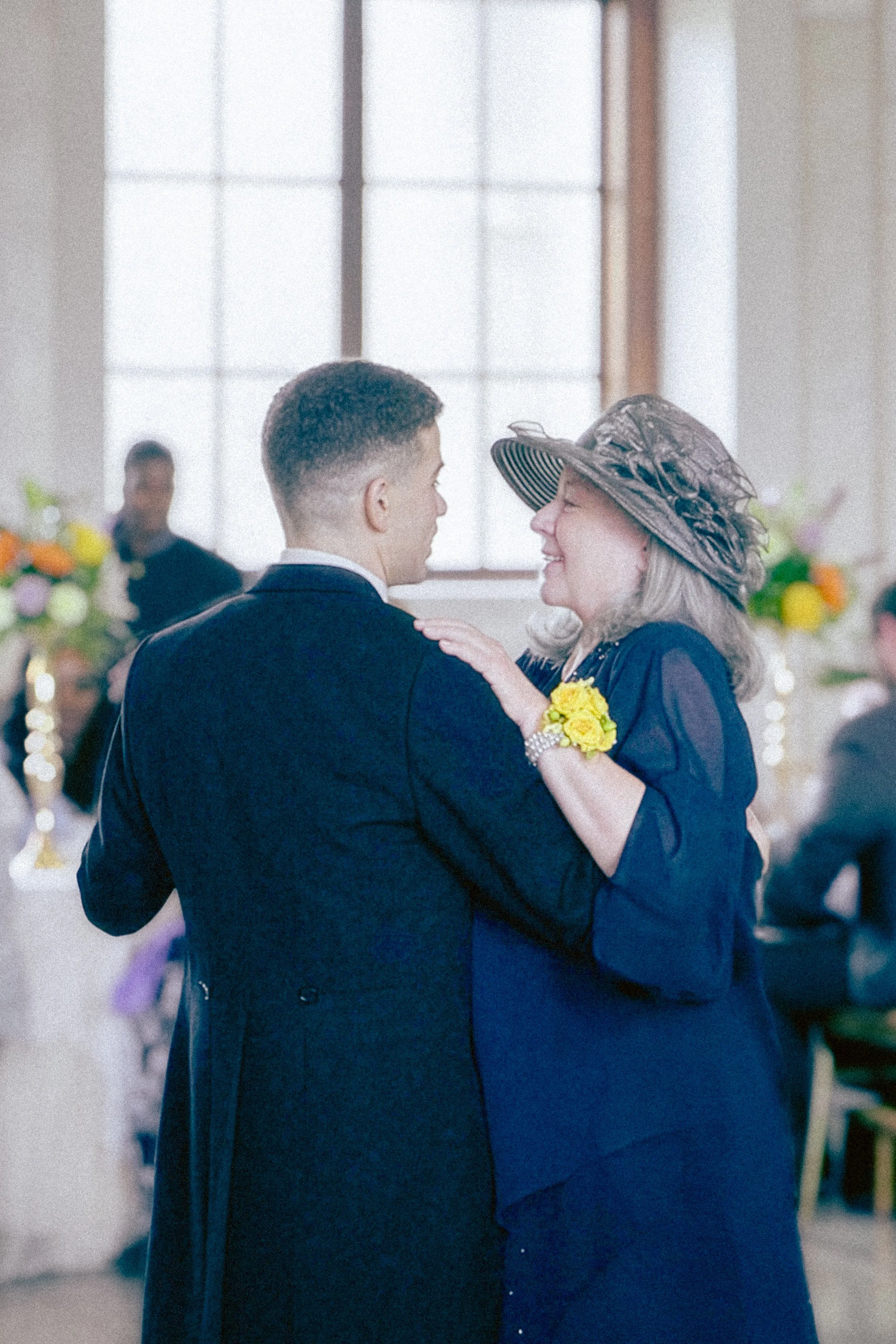 A couple dances closely together during a celebration or wedding reception, with the woman wearing a large floral hat and a dark dress, and the man in a dark suit, inside a decorated venue with large windows and, blurred, colorful floral arrangements