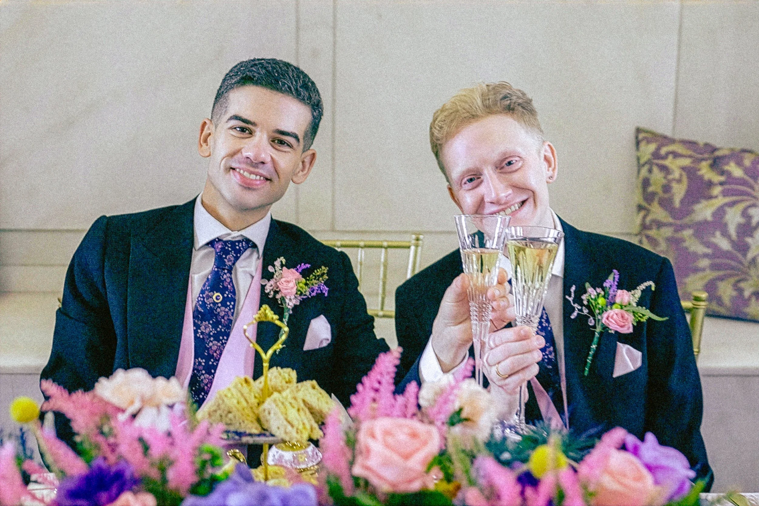 Two men in tuxedos with floral boutonnieres celebrating at a wedding reception, toasting with champagne glasses, surrounded by a colorful flower arrangement.