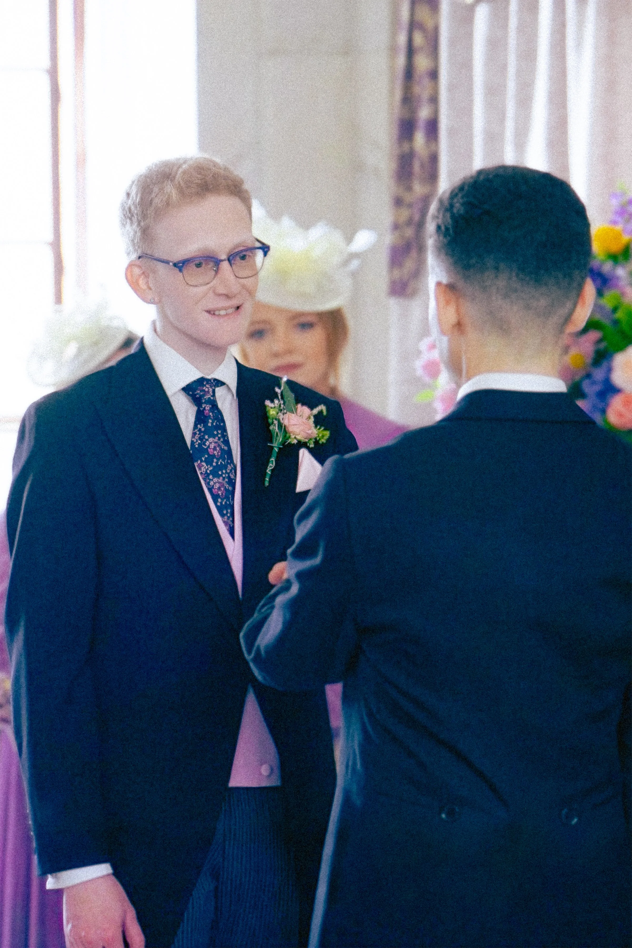 A wedding ceremony with two men in suits exchanging vows, with a woman in a hat and floral dress present in the background, decorated with flowers and curtains.