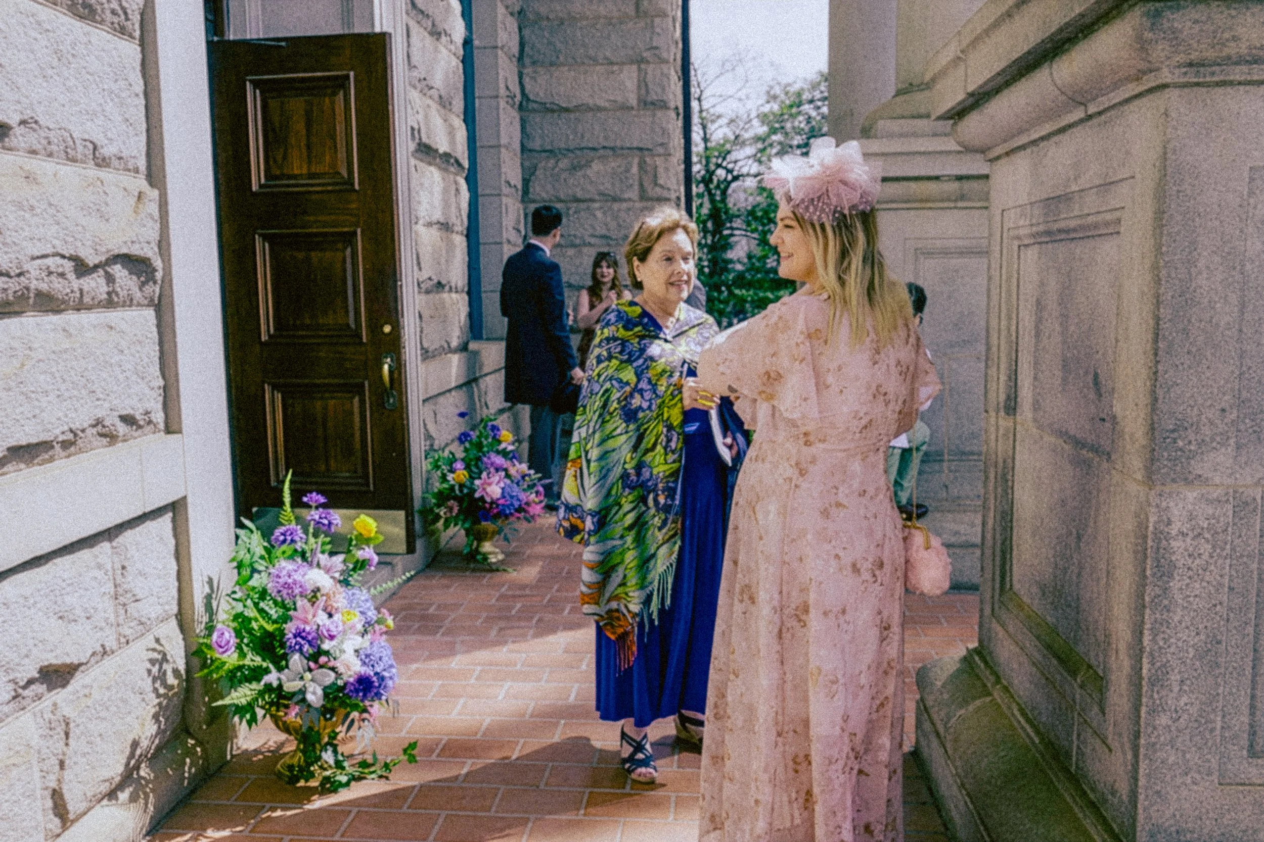 People talking outside a building entrance decorated with flowers, including two women in colorful dresses and a pink hat, with a man and woman in the background.
