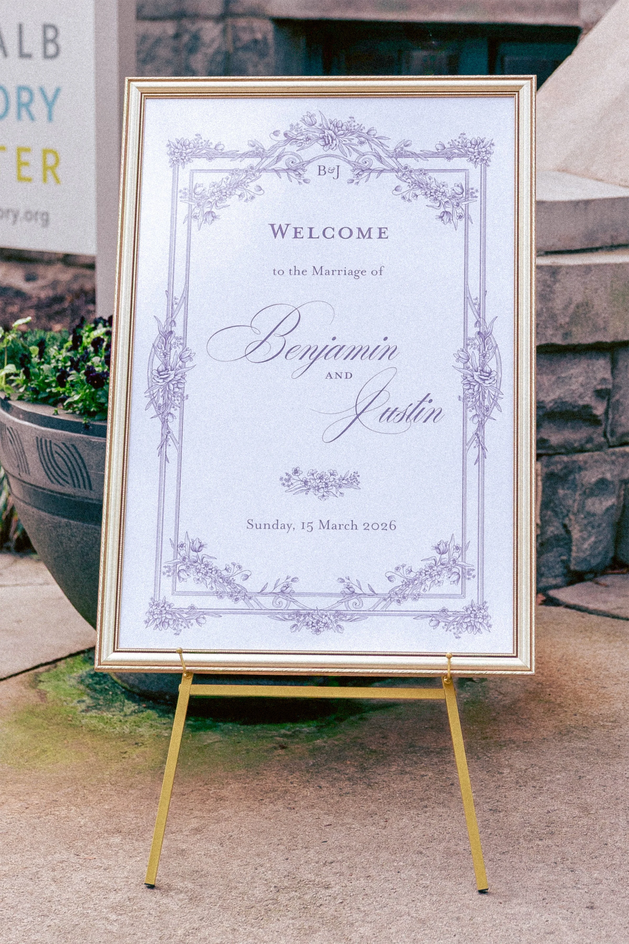 A wedding welcome sign with elegant purple floral border and text, welcoming guests to the marriage of Benjamin and Austin on Sunday, 15 March 2026. The sign is displayed on an easel outside a building with stone stairs and a large black planter with