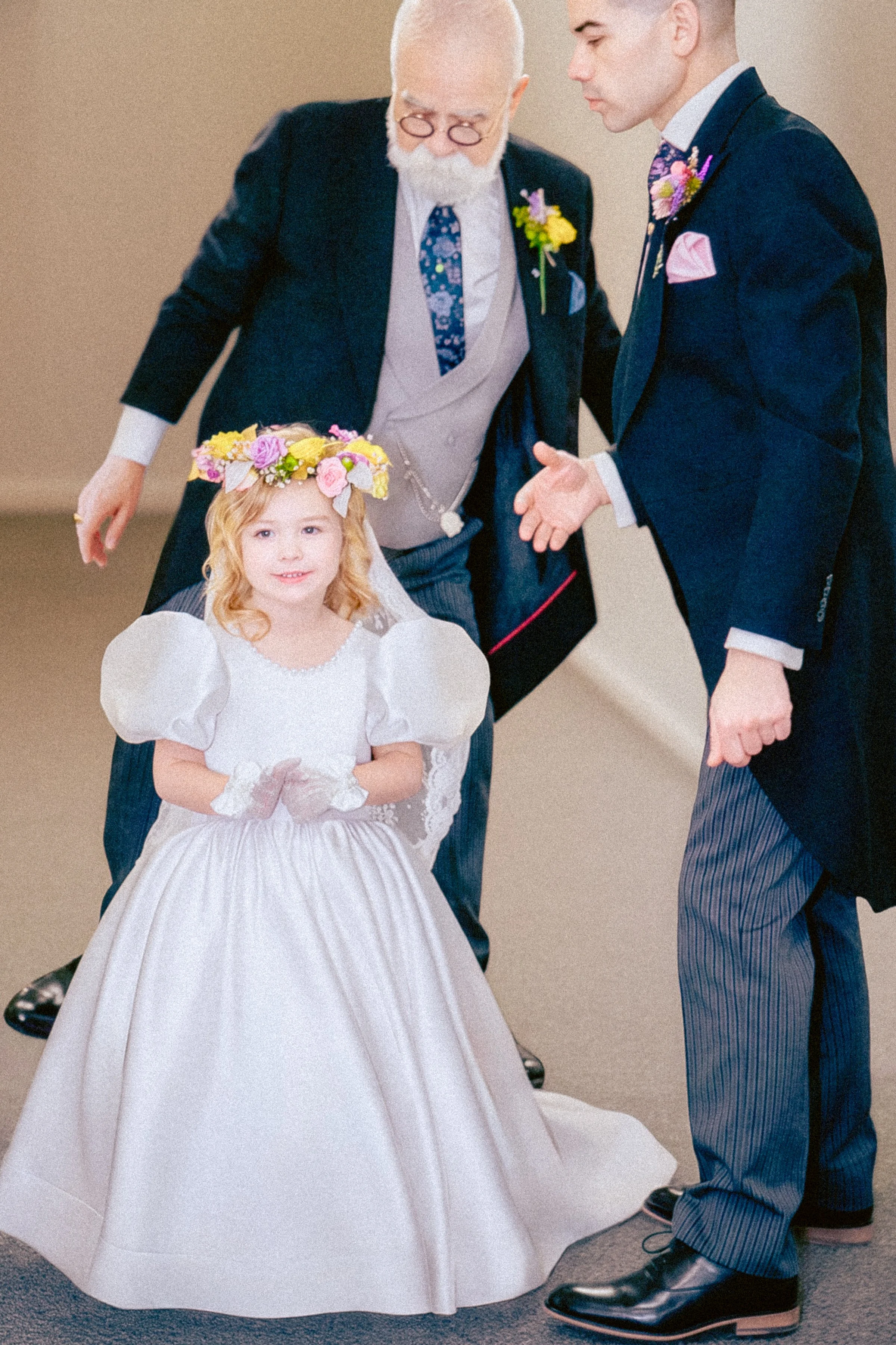A young girl in a white dress and floral crown standing with two men in suits at a formal event.