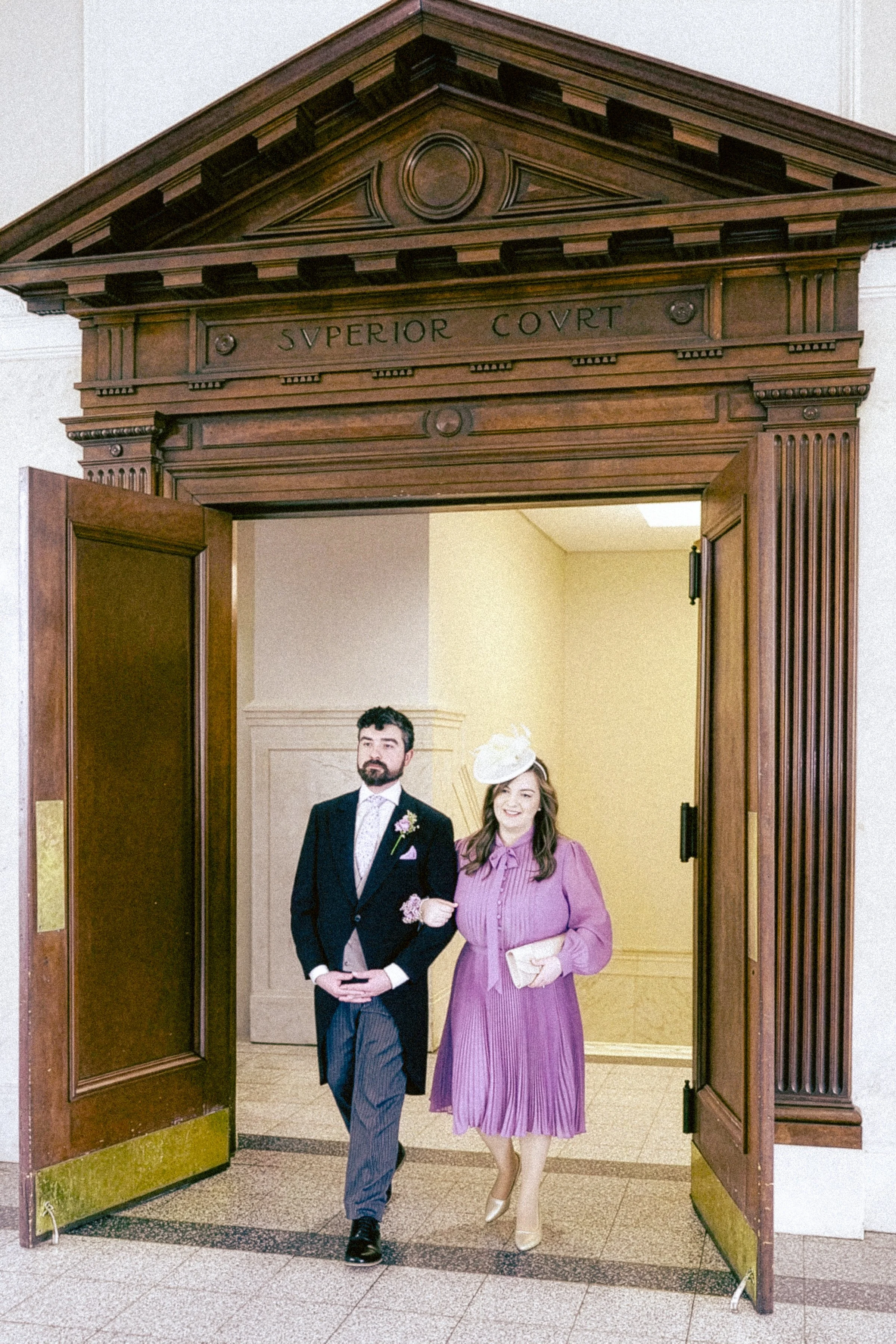 A man and a woman walking through a doorway. The man is wearing a formal suit, and the woman is dressed in a pink vintage style dress with a matching hat and clutch purse. The doorway has wooden framing with the inscription 'SVPERIOR COVRT' at the to