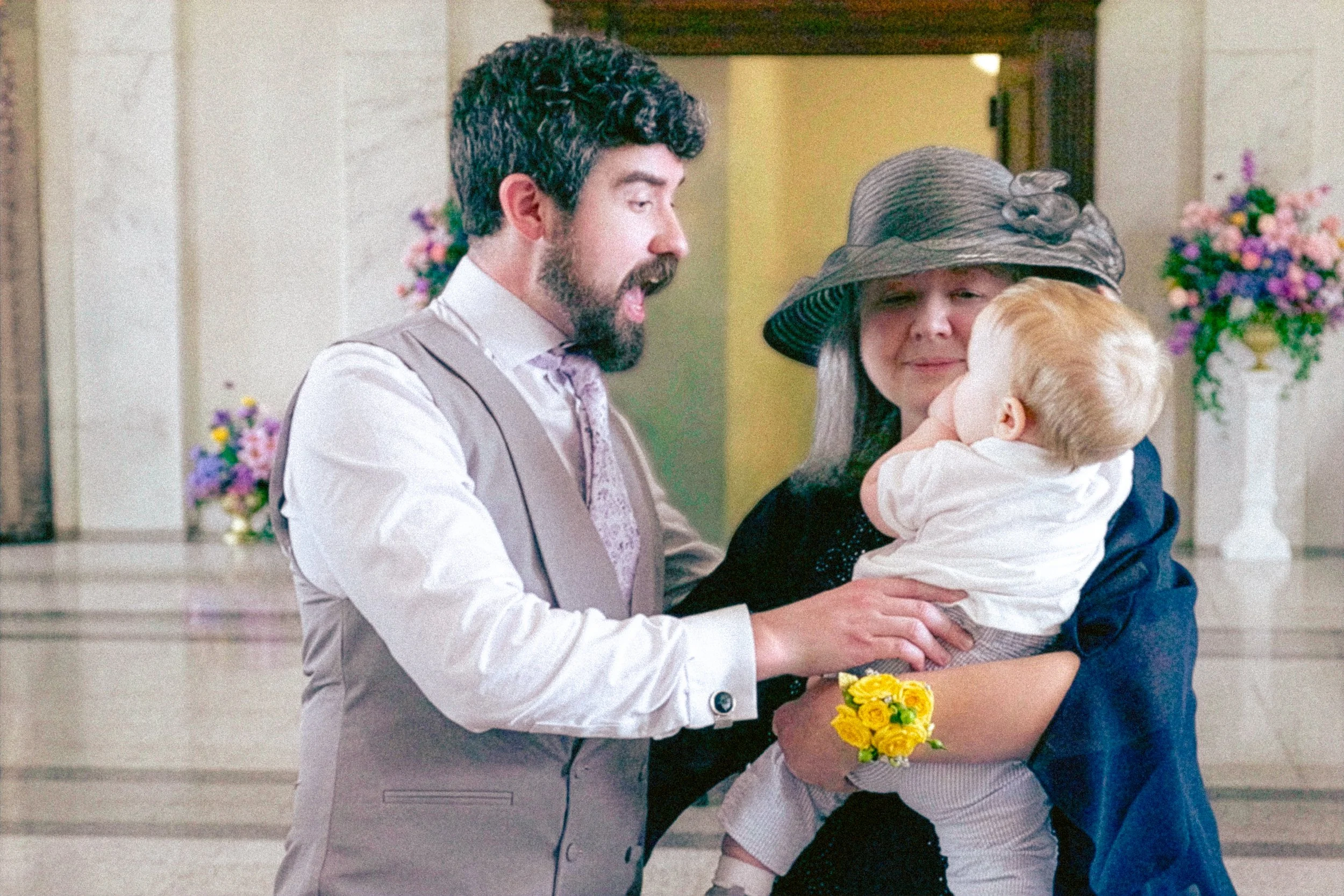A man in a beige vest and tie, a woman in a large hat, and a child in white shirt, sharing a moment at a wedding with flowers in the background.