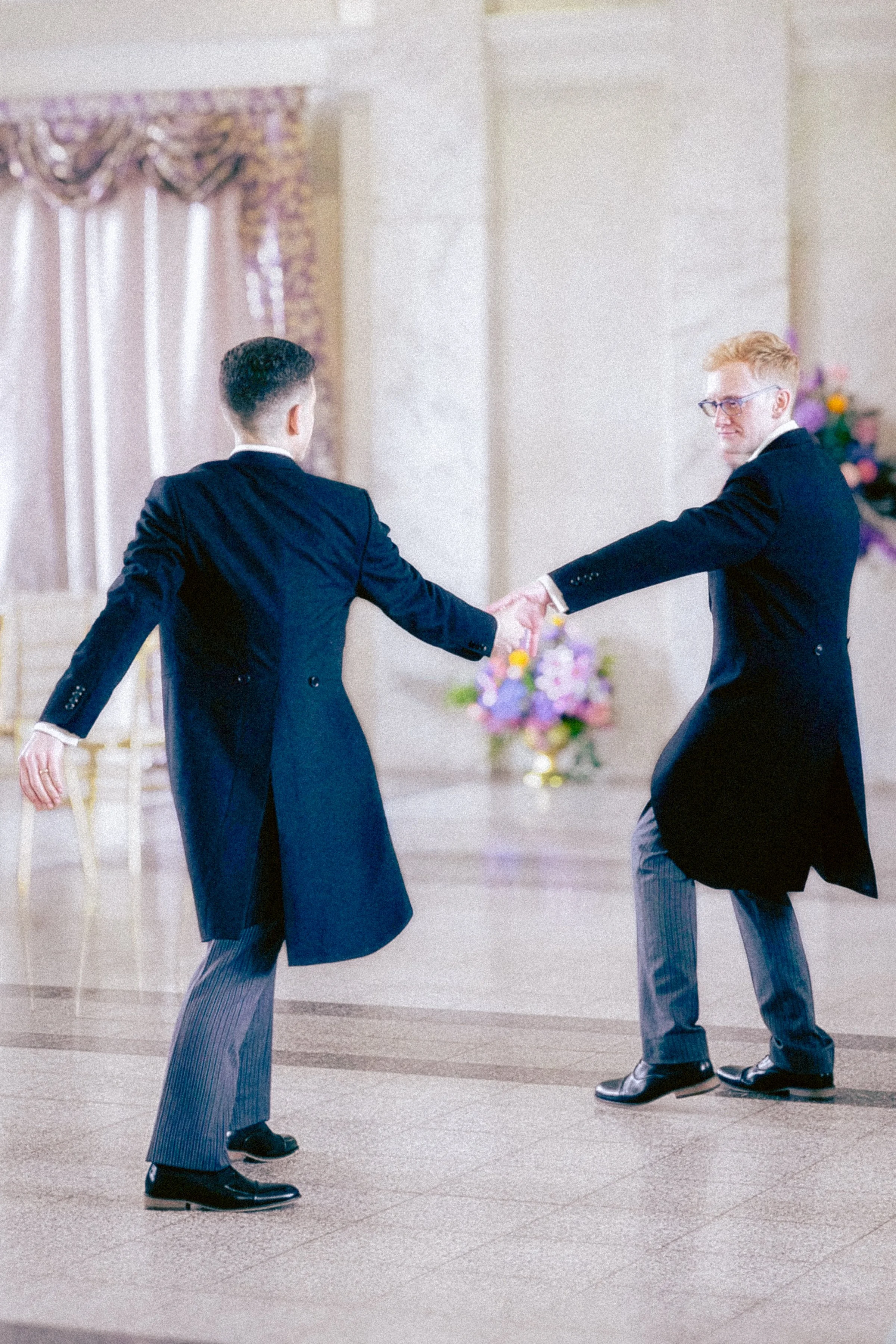 Two men in formal suits holding hands and dancing in a banquet hall with floral decorations.