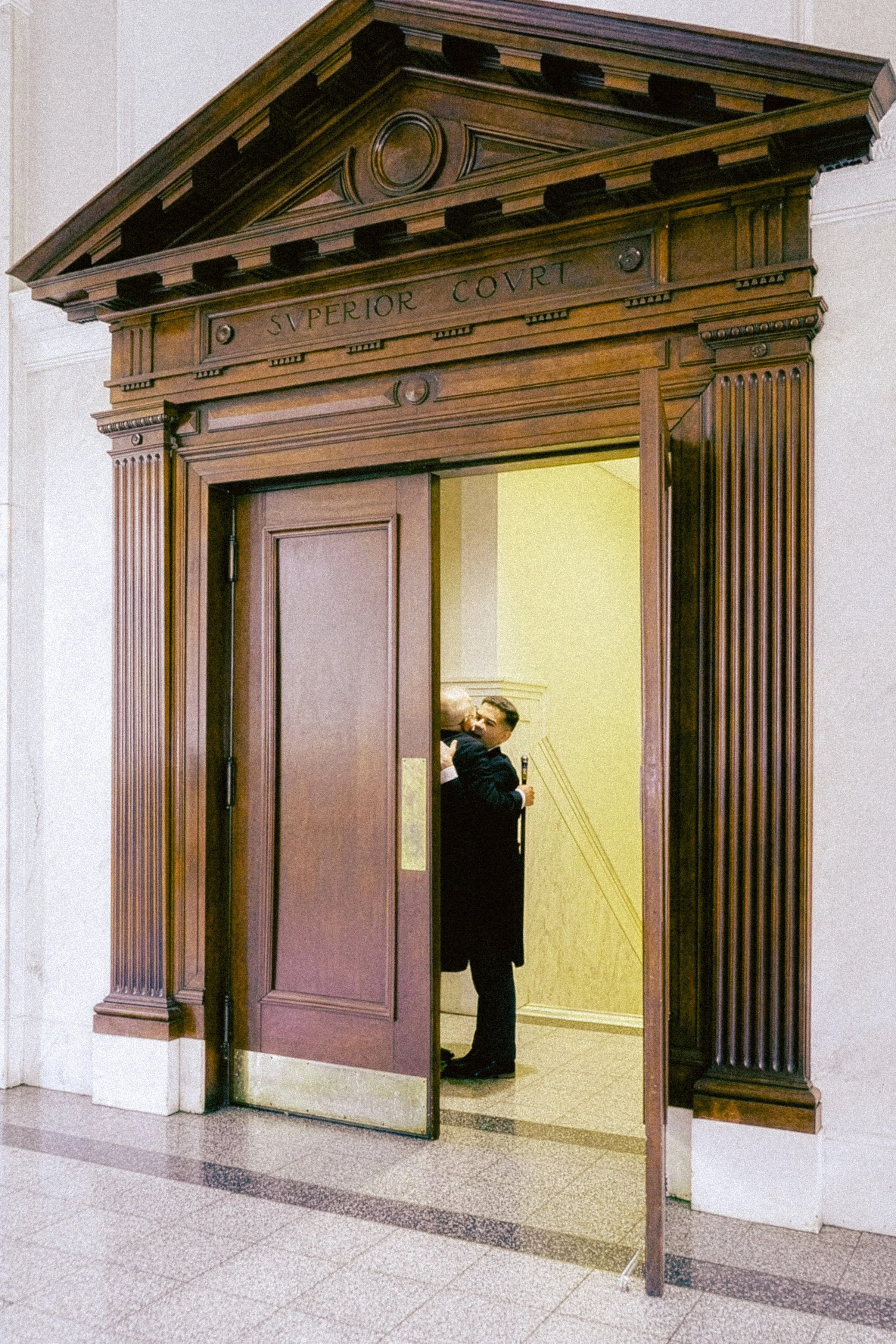 Wooden doorway with a triangular pediment labeled "Superior Court". A person standing inside, held a drink, is visible through the open door.