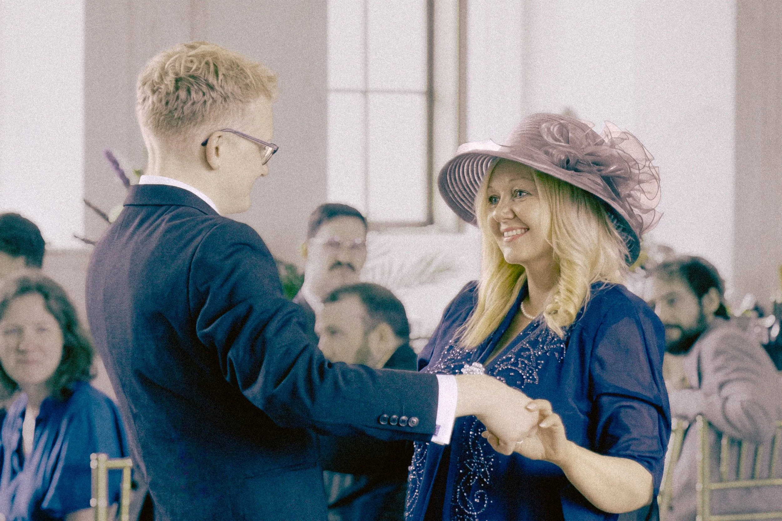 A young man and woman in formal attire dancing together at a wedding or similar celebration, surrounded by seated guests in a decorated hall.