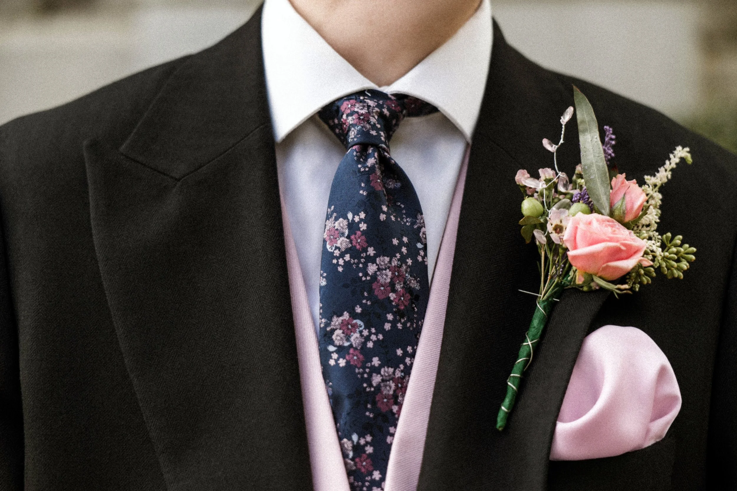 Close-up of a man wearing a dark suit, white shirt, floral patterned tie, and a boutonniere with pink roses and purple flowers on the lapel.