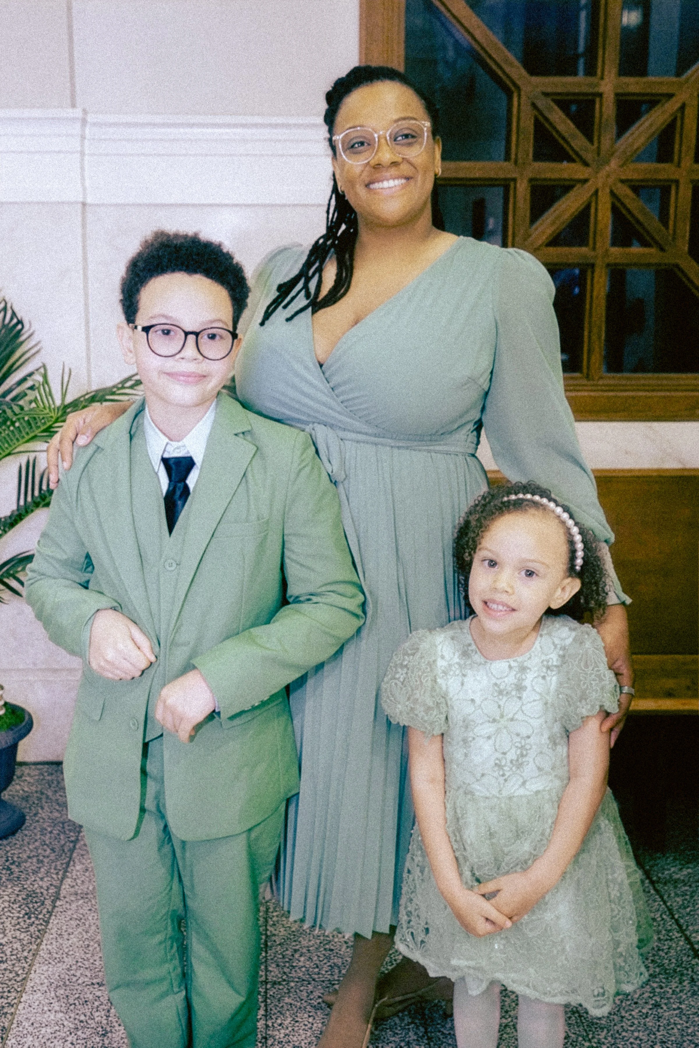 A group of four people standing indoors, smiling at the camera. Two children and two adults. The boy and girl are dressed up in formal attire, with the boy in a green suit and glasses, and the girl in a lace dress and hair accessories. One adult woma