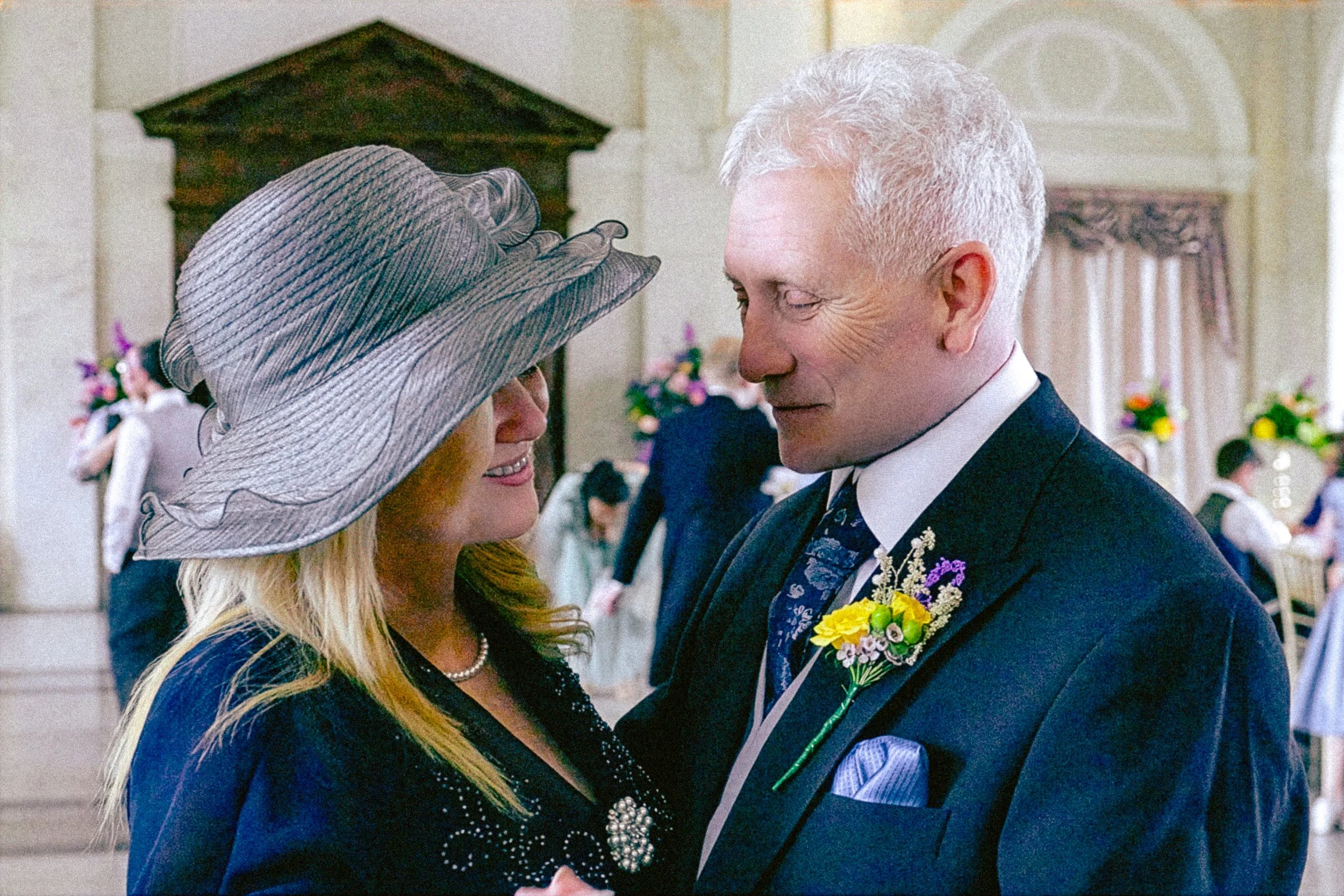 A couple dancing closely, dressed in formal attire, with the woman wearing a large hat with ribbons and the man in a tuxedo with a floral boutonniere, indoors at a decorated event.