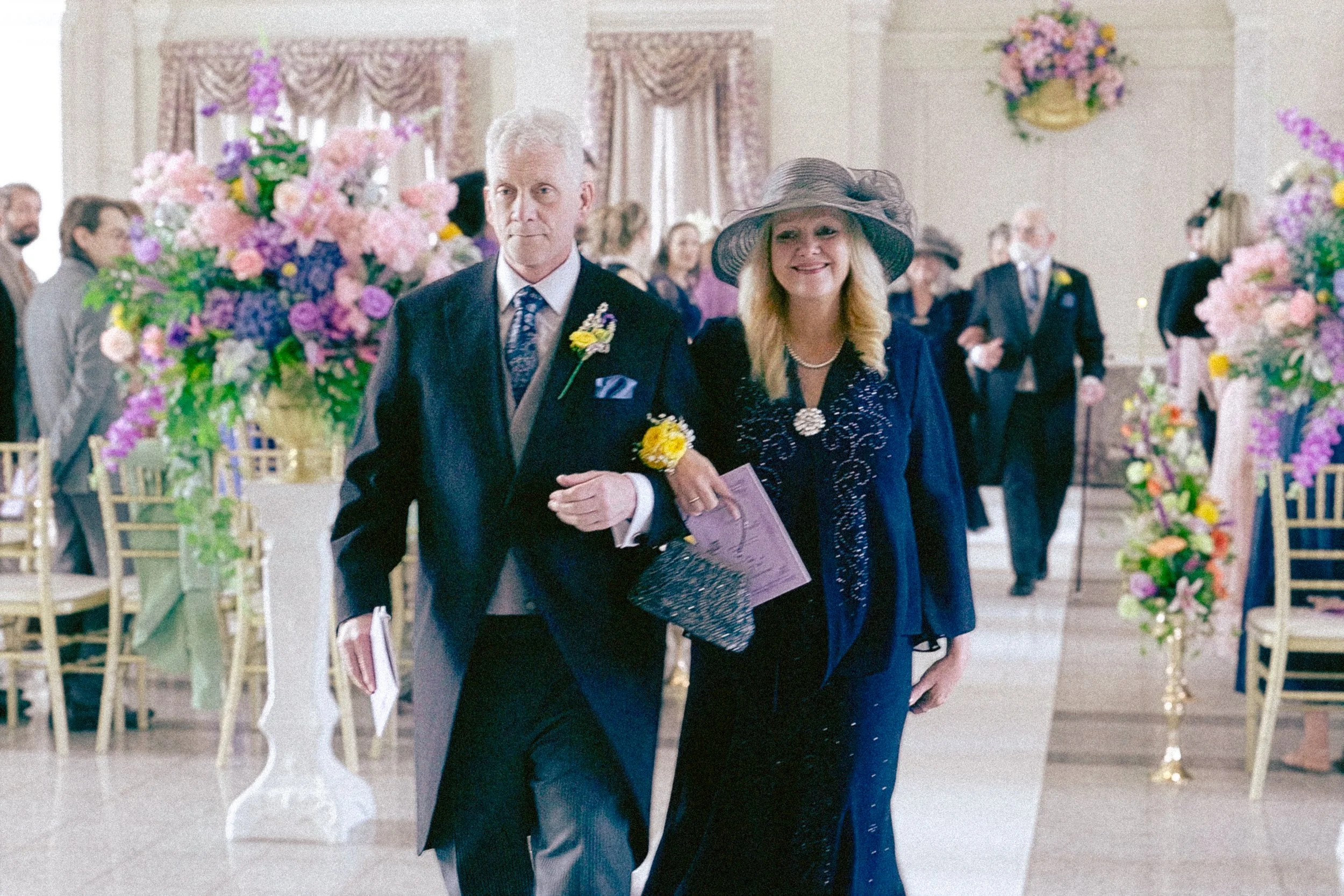 A couple dressed in formal attire walking hand in hand at a wedding reception. The man is wearing a dark suit with a boutonnière, and the woman is wearing a dark dress with a wide-brimmed hat. The background shows elegant floral arrangements and othe