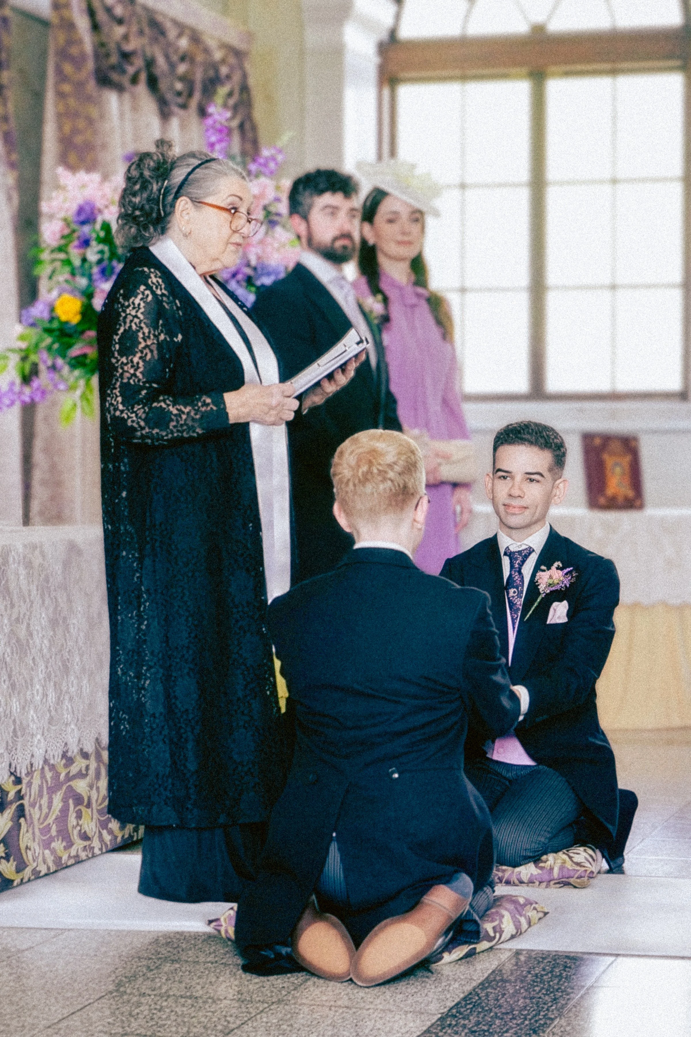 A wedding ceremony with a priest officiating, two men kneeling before each other, and a woman standing in the background near floral decorations and a window.