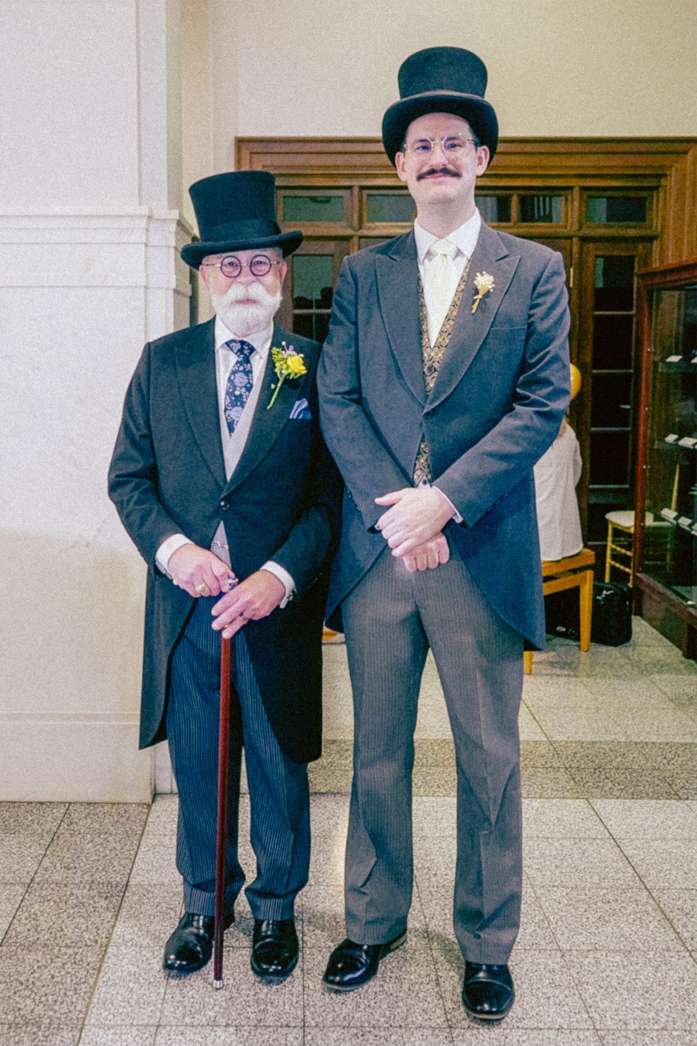Two men dressed in vintage formal attire, each wearing a tall top hat. The man on the left has a white beard and glasses, holding a cane, and wears a dark suit with a floral tie and a yellow boutonniere. The man on the right has a mustache and glasse