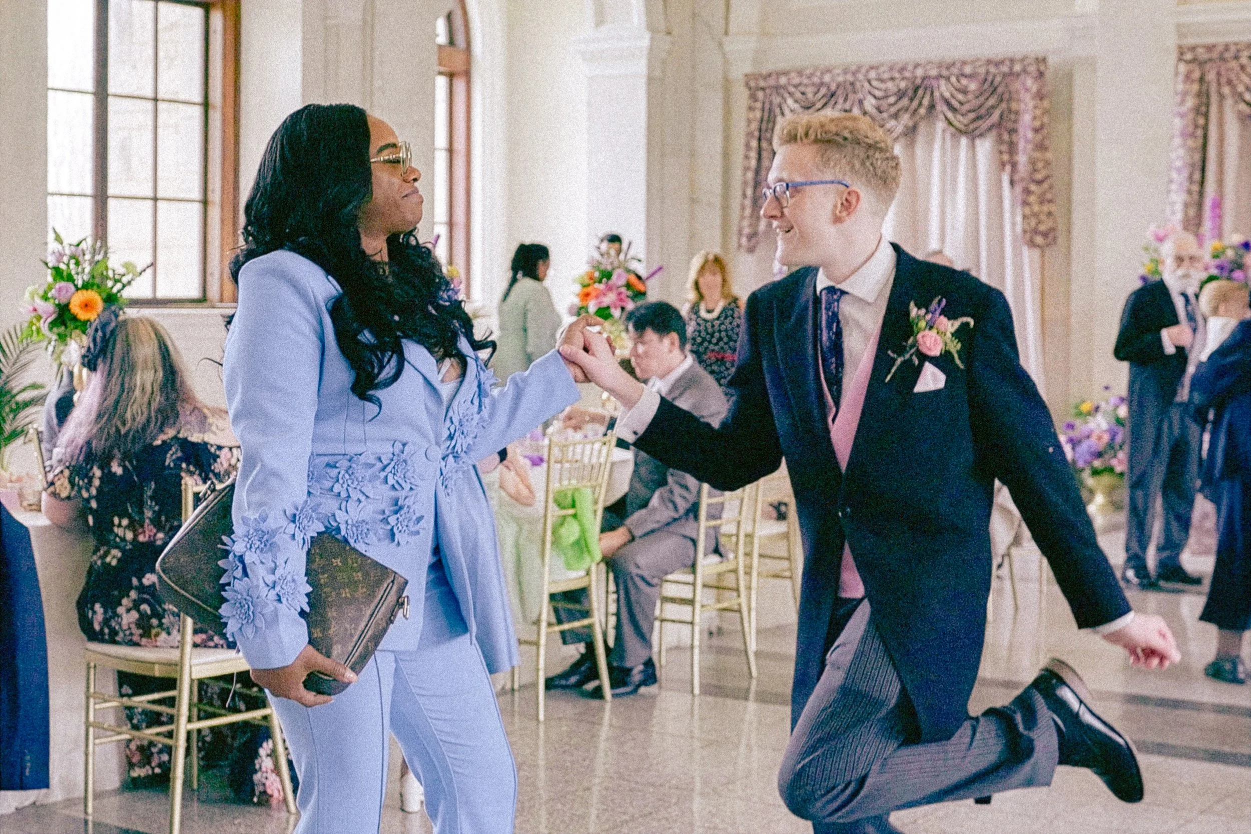 A man and woman are holding hands and dancing at a wedding reception in a decorated hall with floral arrangements.