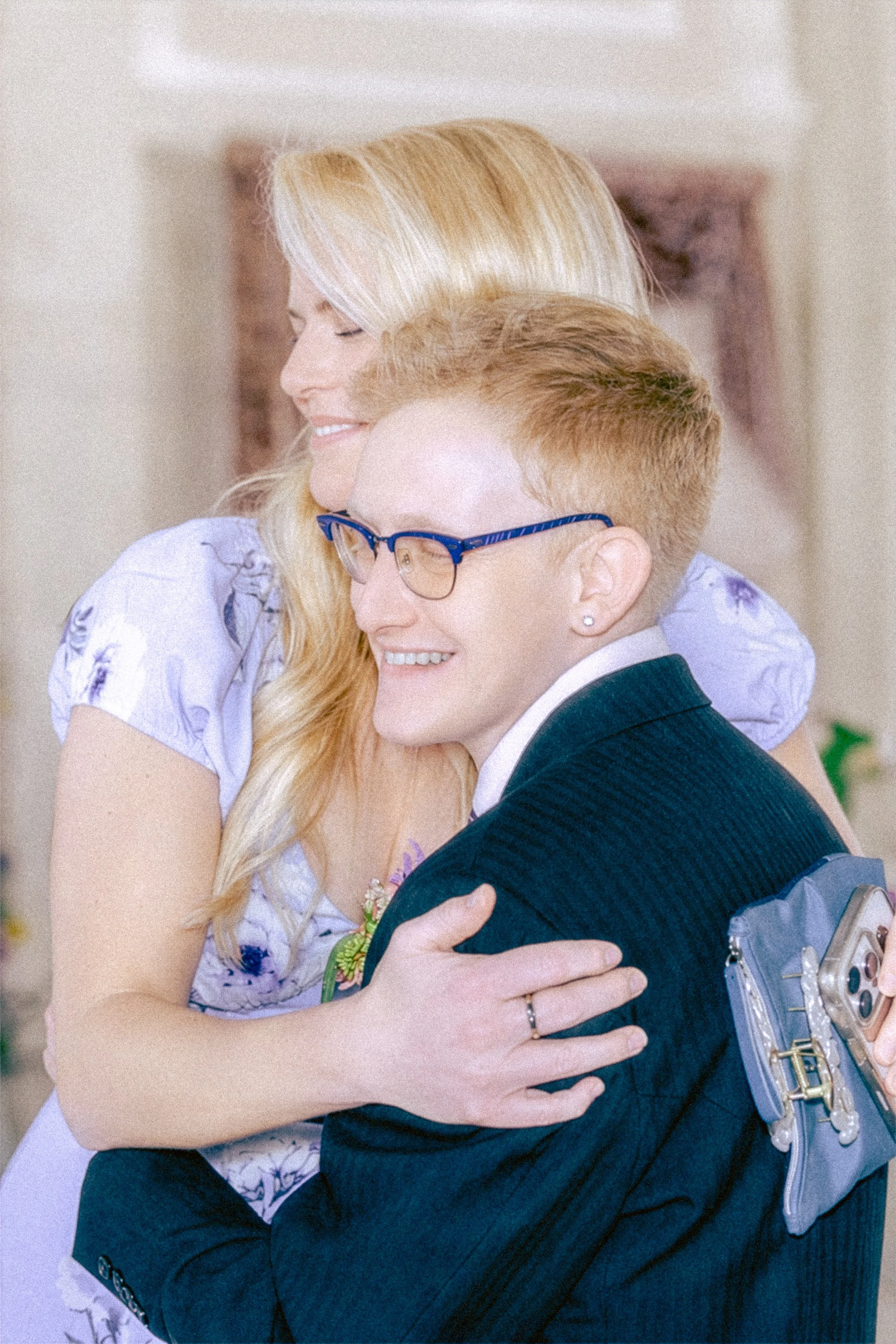 A woman with long blonde hair is hugging a man with short blonde hair and glasses. The woman has her eyes closed and is smiling, wearing a white dress with floral patterns. The man, dressed in a dark suit, is smiling and wearing a futuristic vest wit