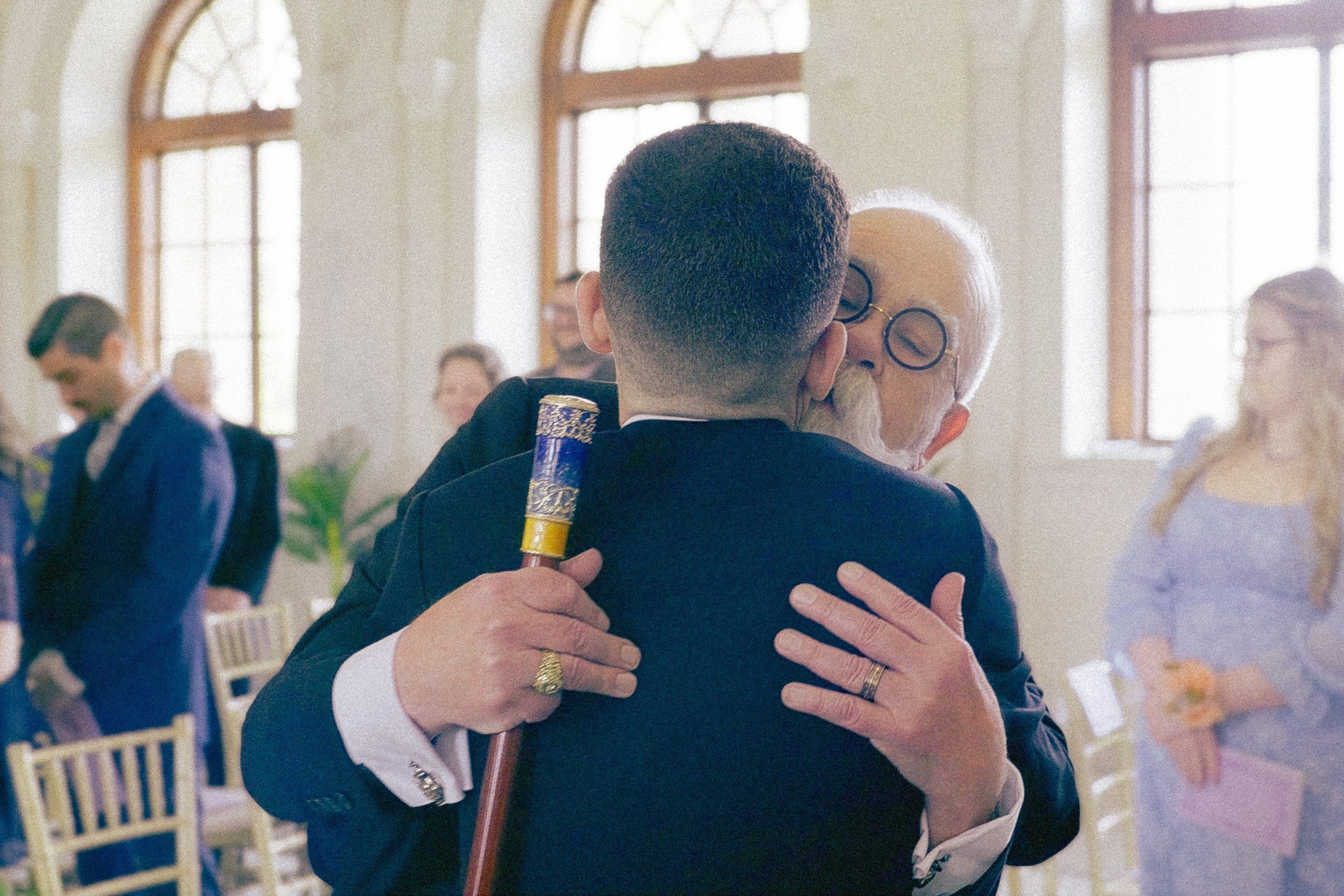 A man and an elderly woman are hugging in a bright room with large windows, with other people in the background.